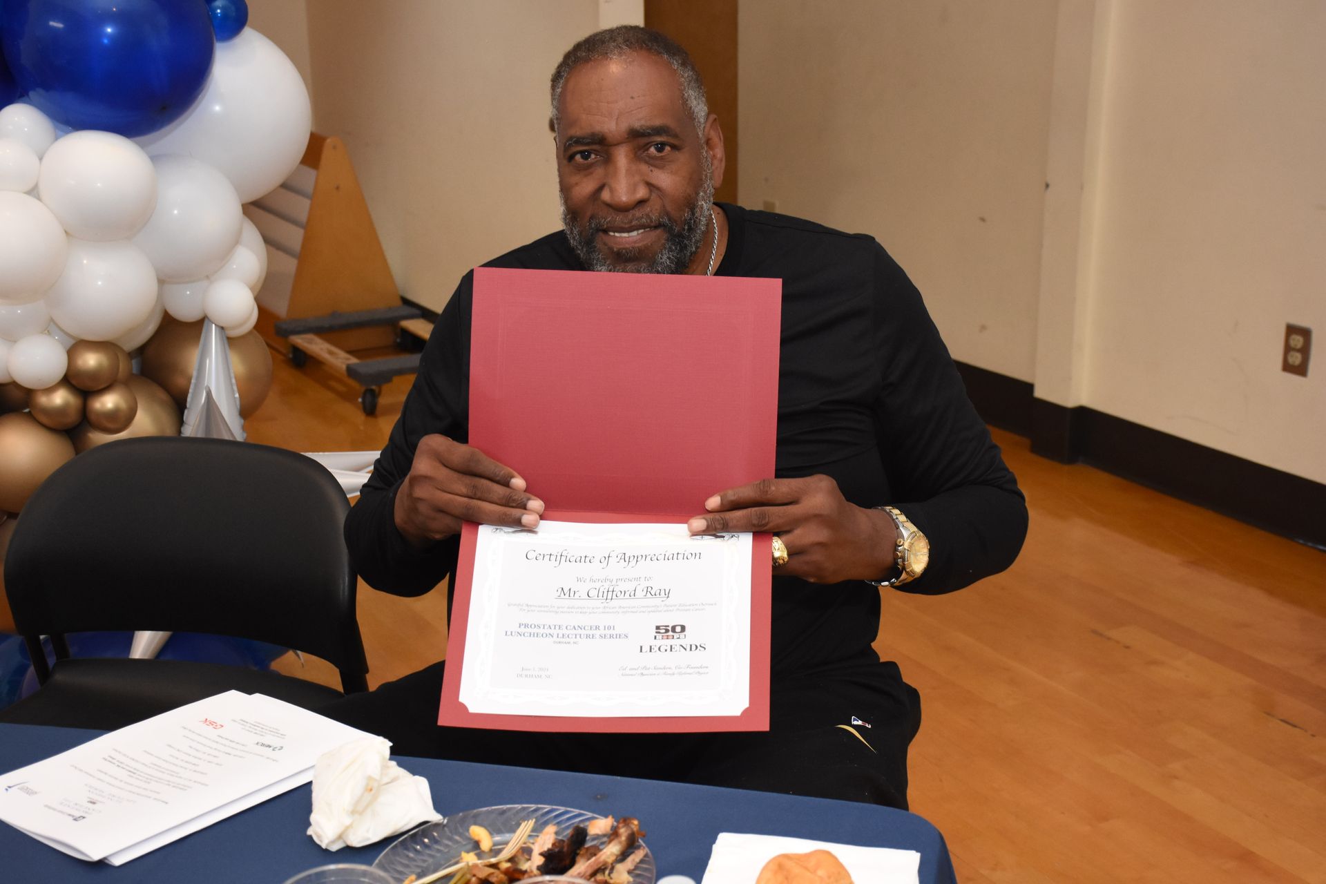 Man holding a certificate, smiling at a celebratory event; balloons in background.