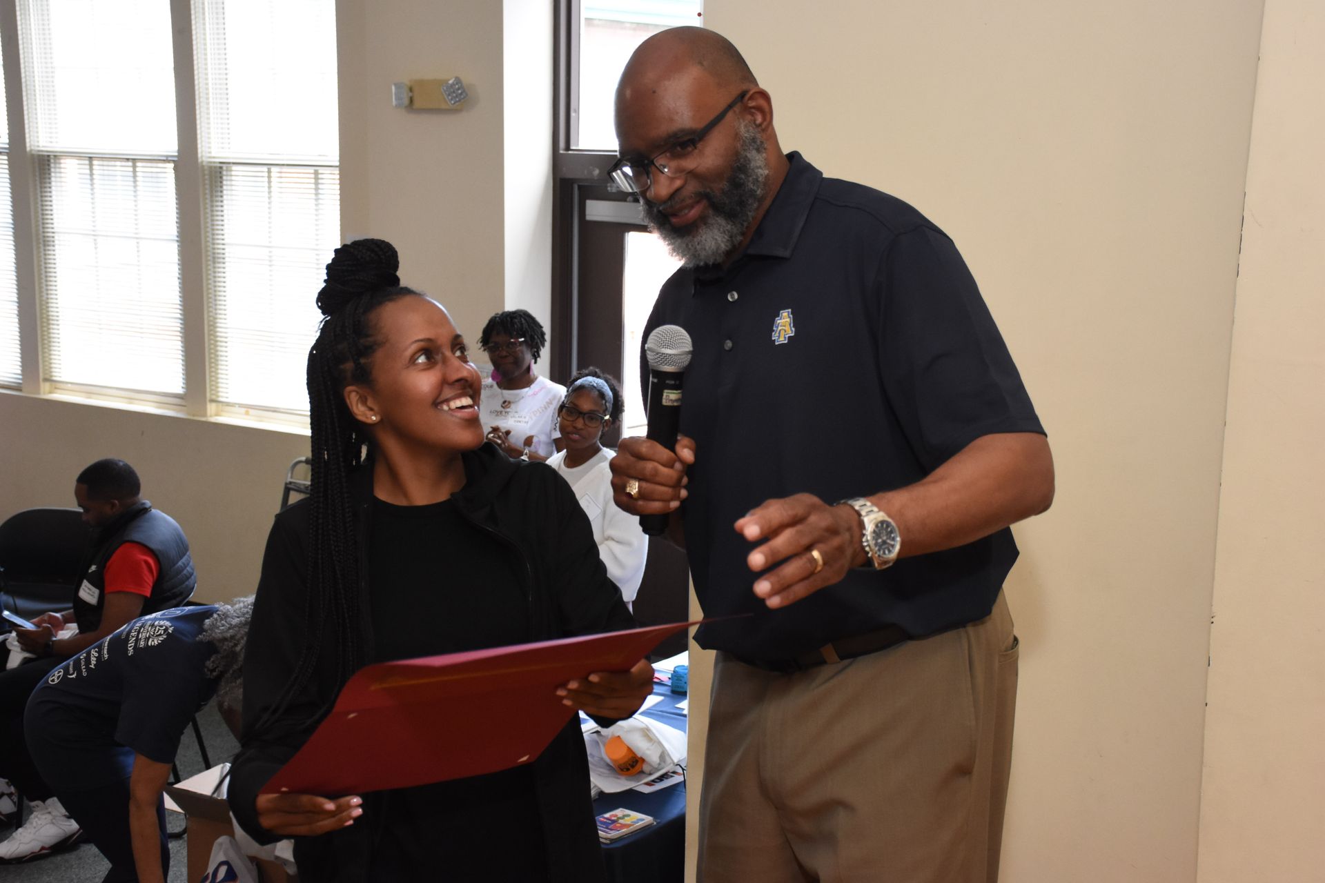 Man presents a certificate to a woman in a room, both smiling. The man holds a microphone.