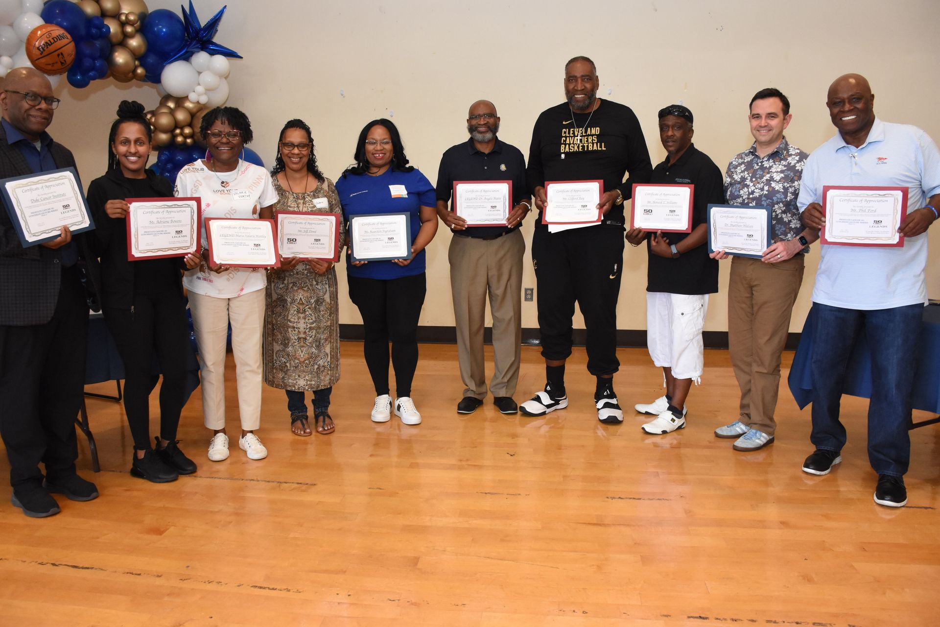 Group of people holding framed certificates, possibly at an award ceremony. Balloons in background.