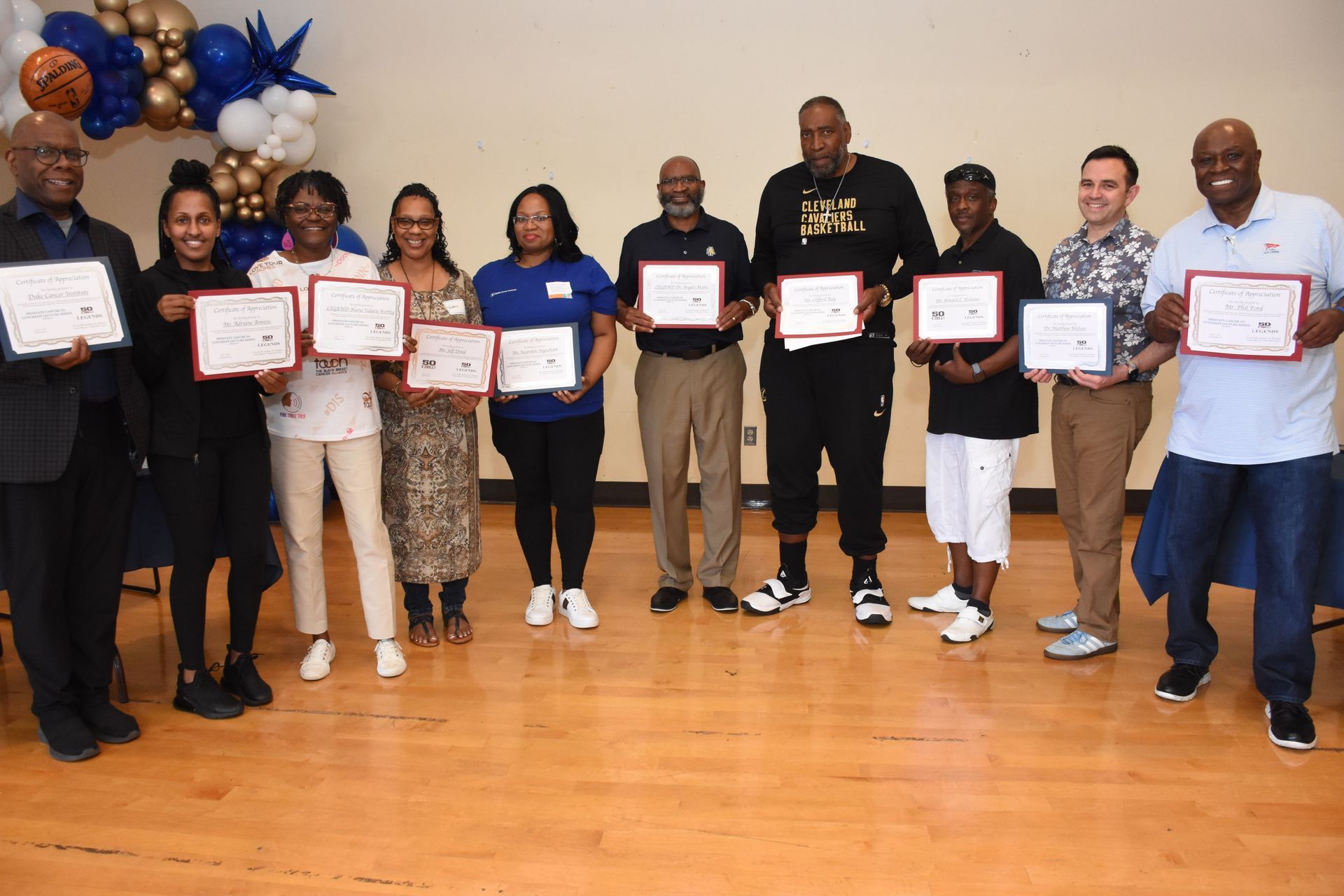 Group of people holding certificates, smiles for the camera, balloons in background, indoor setting.