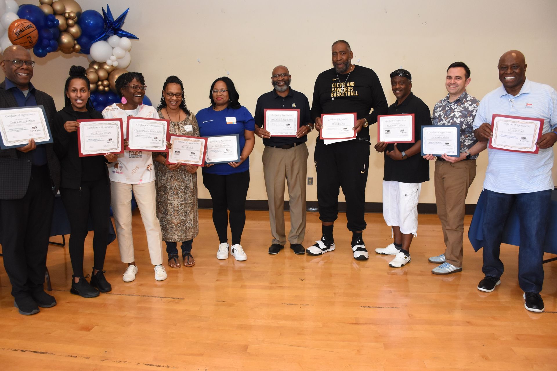 Group of people holding certificates, smiling, indoors. Balloons in background.