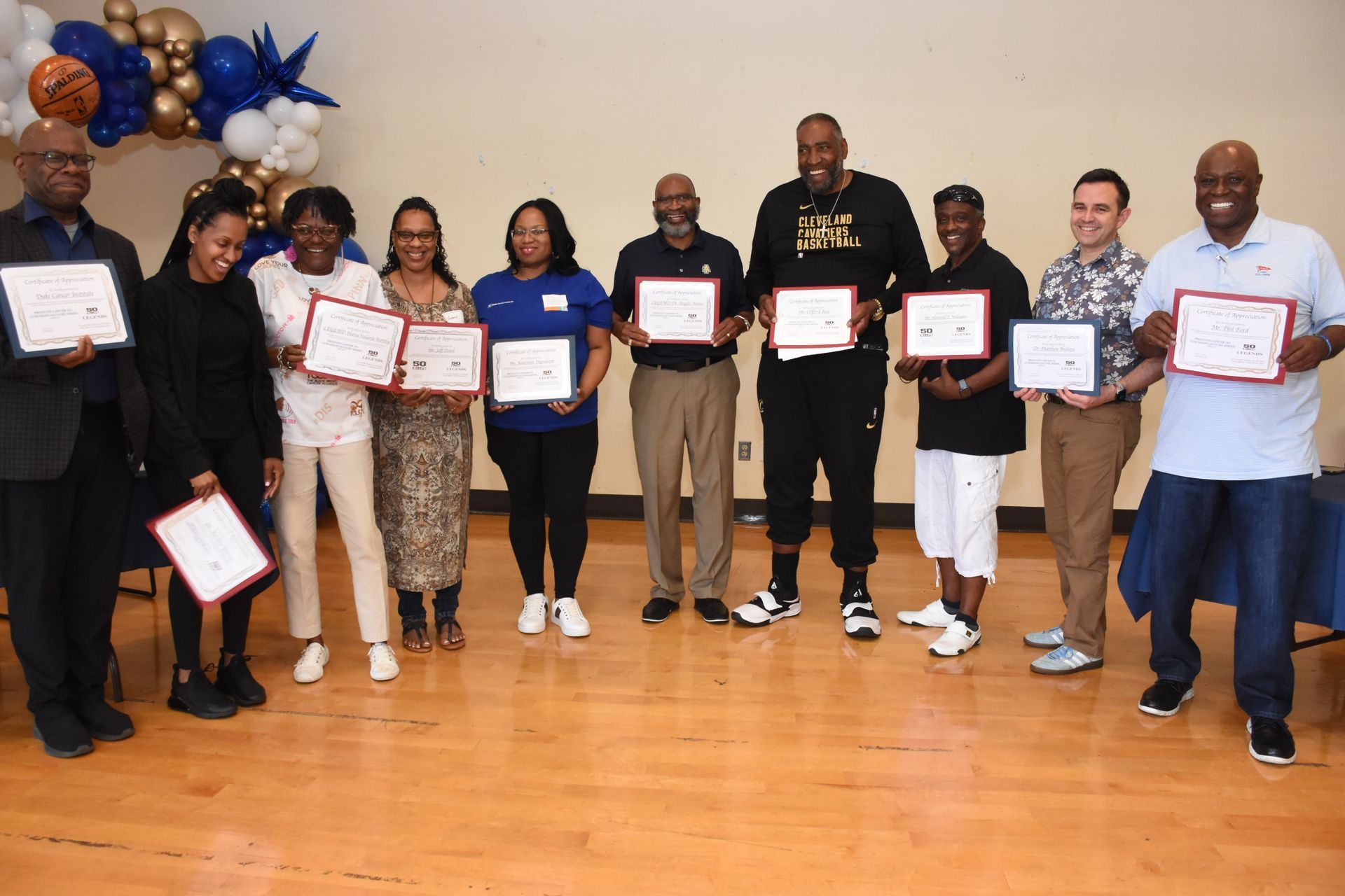 Group of people holding certificates, smiling, indoors. Balloons in background.