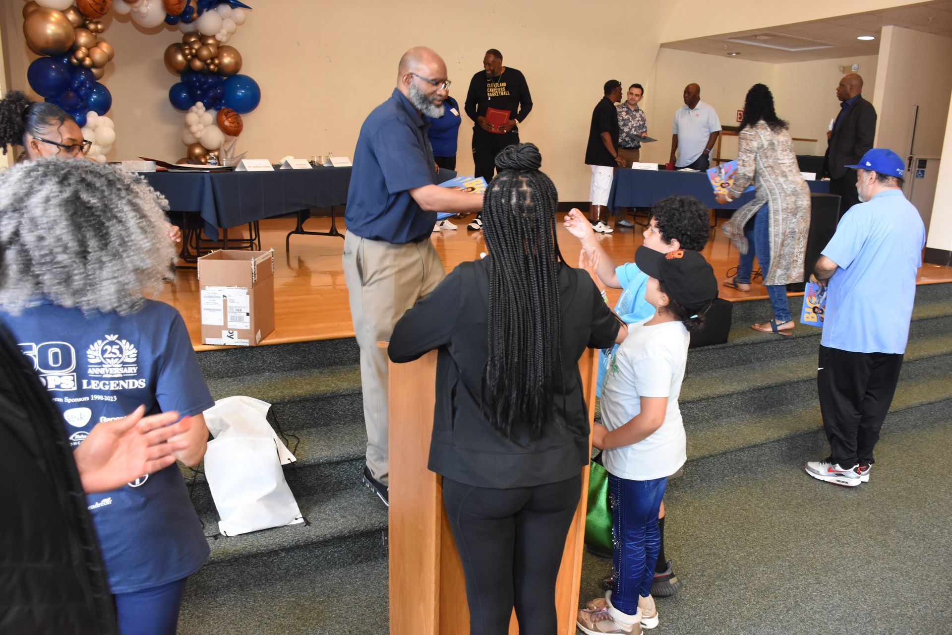 Man handing out items to children at an event. Balloons and tables in background.