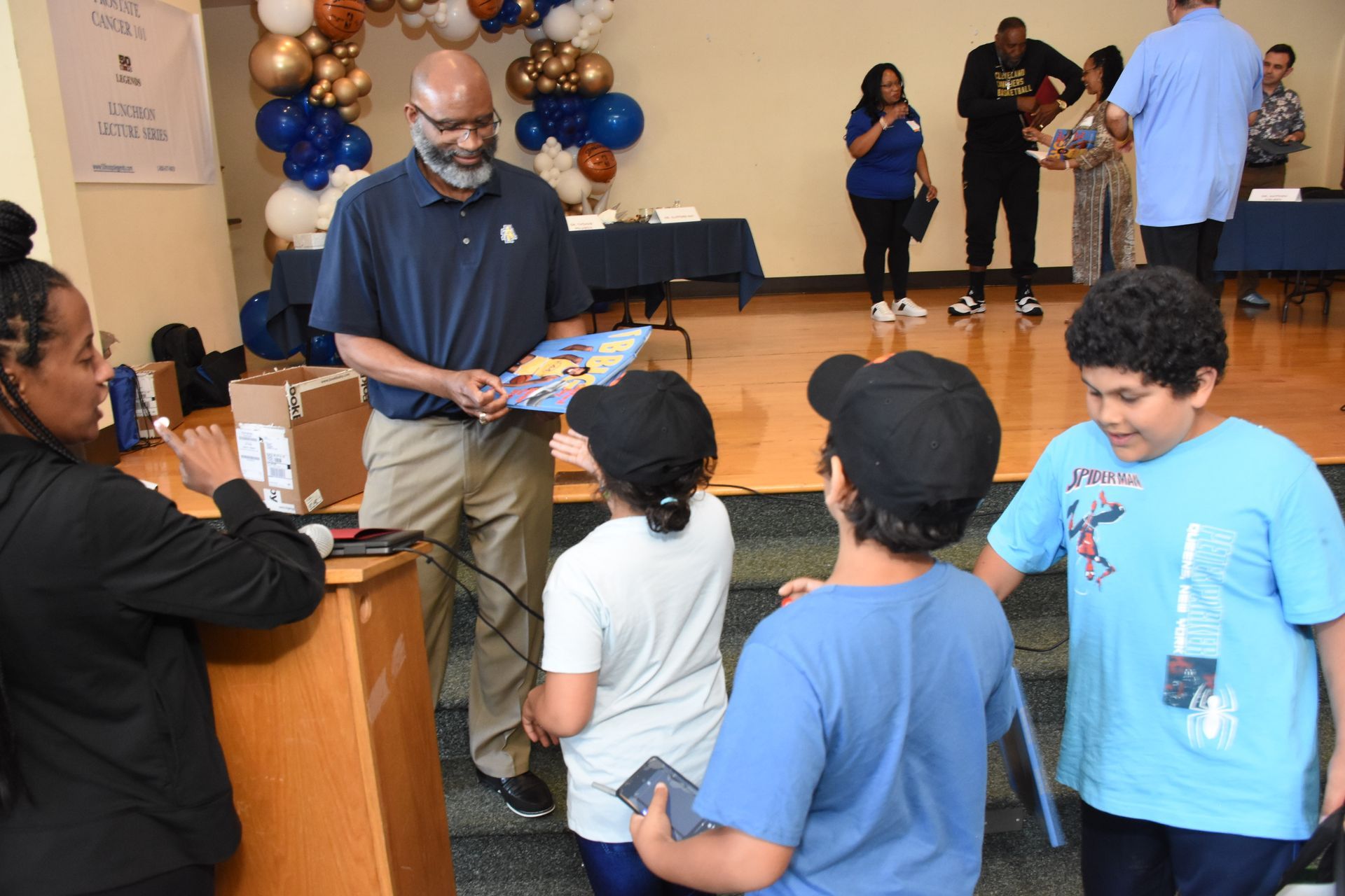 Man handing a book to children at a community event. Blue and gold balloons decorate the room.
