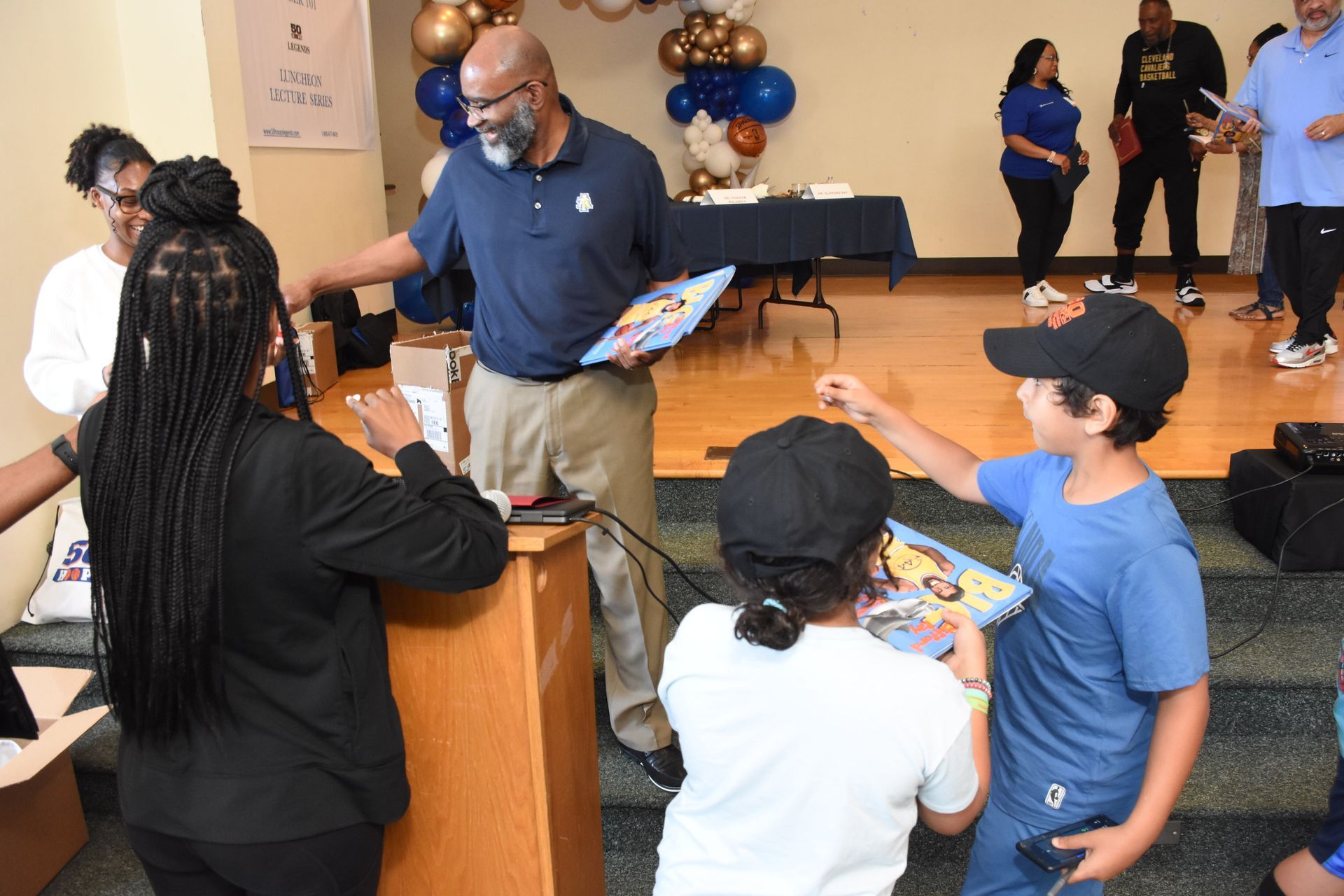 Man handing books to children at an event, with balloons and other people in the background.