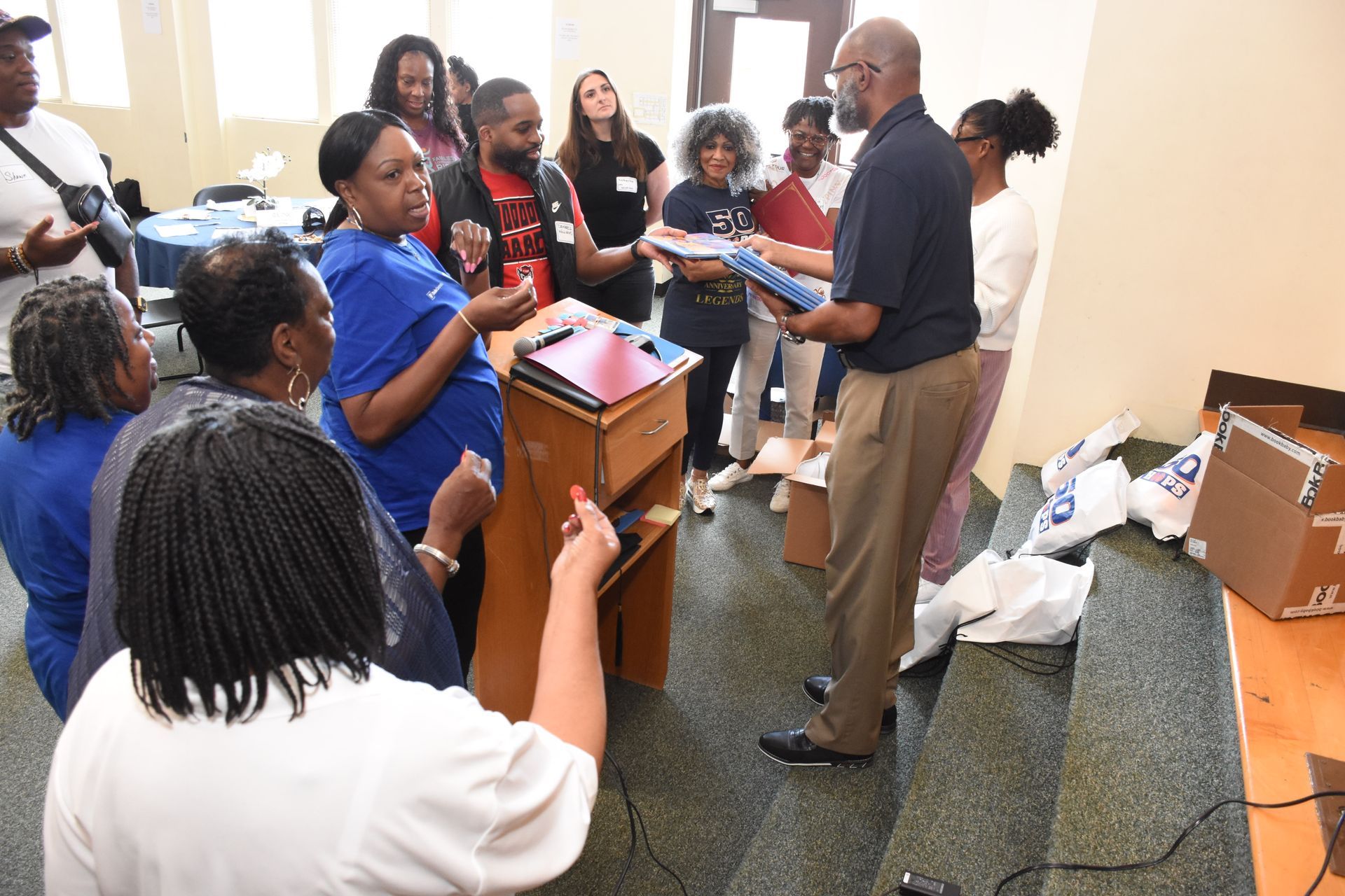 Group of people in a room; a man hands out items. Some wear blue shirts. Others watch, some near a podium.