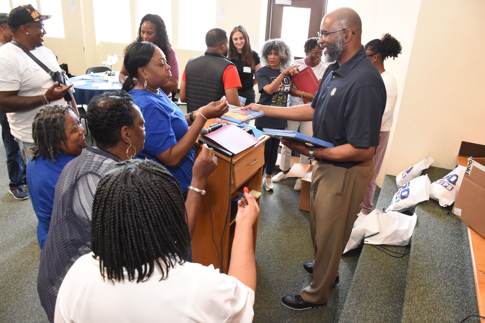 A man hands materials to a group of people. Some wear blue and are gathered around a table.