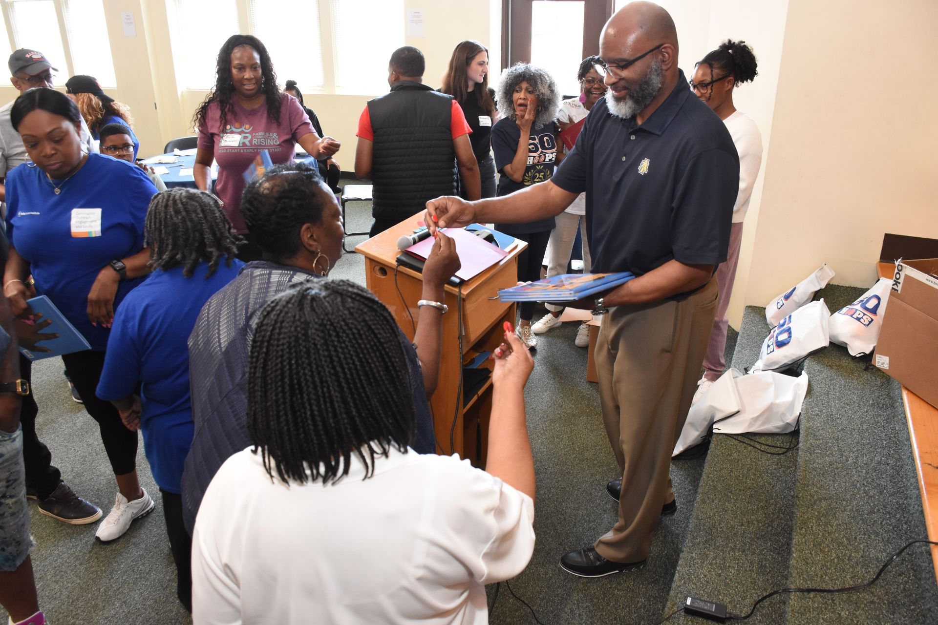 Man handing out materials to a line of people in a room; some are holding blue folders.