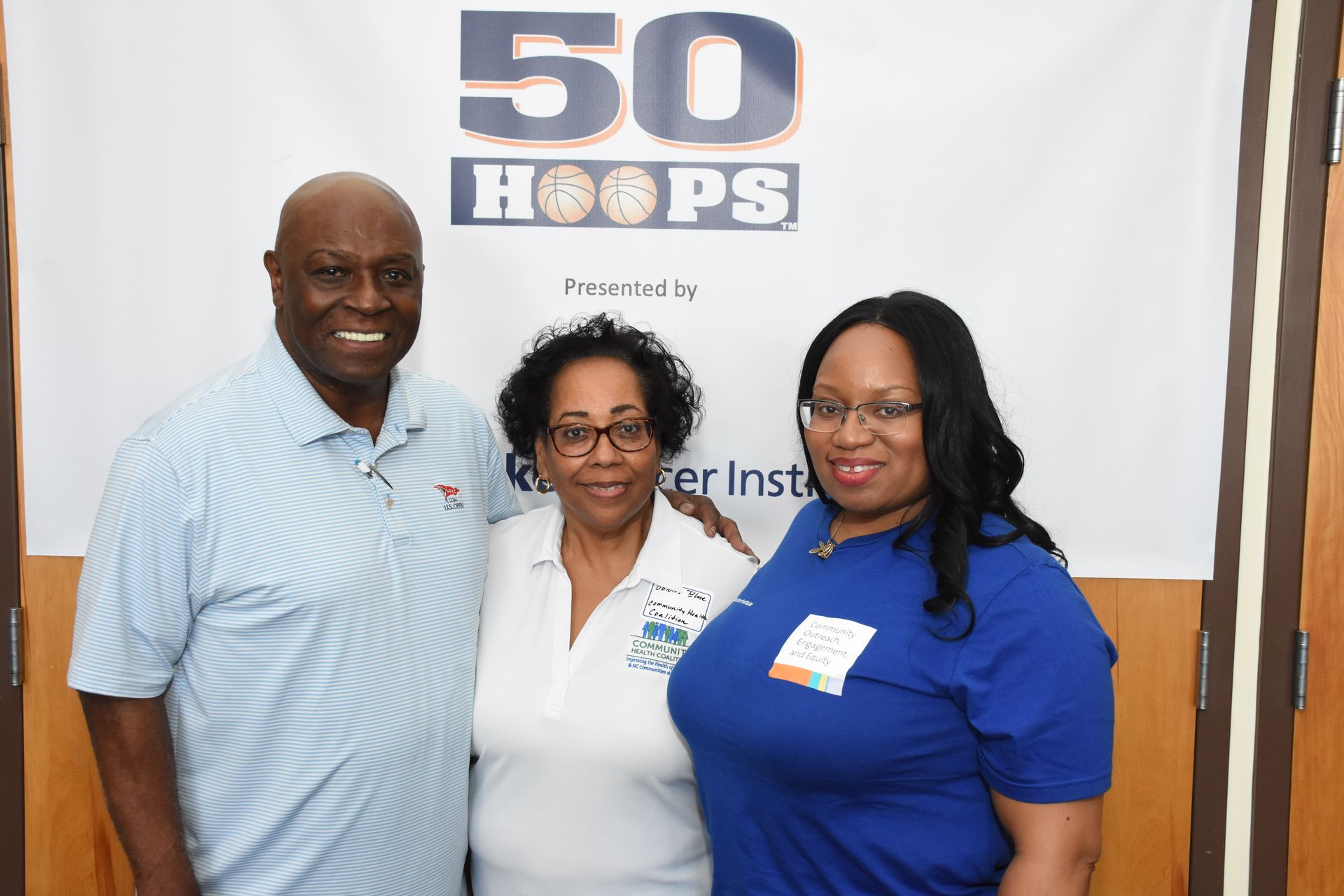 Three people pose in front of a banner. A man in a blue shirt smiles next to two women.