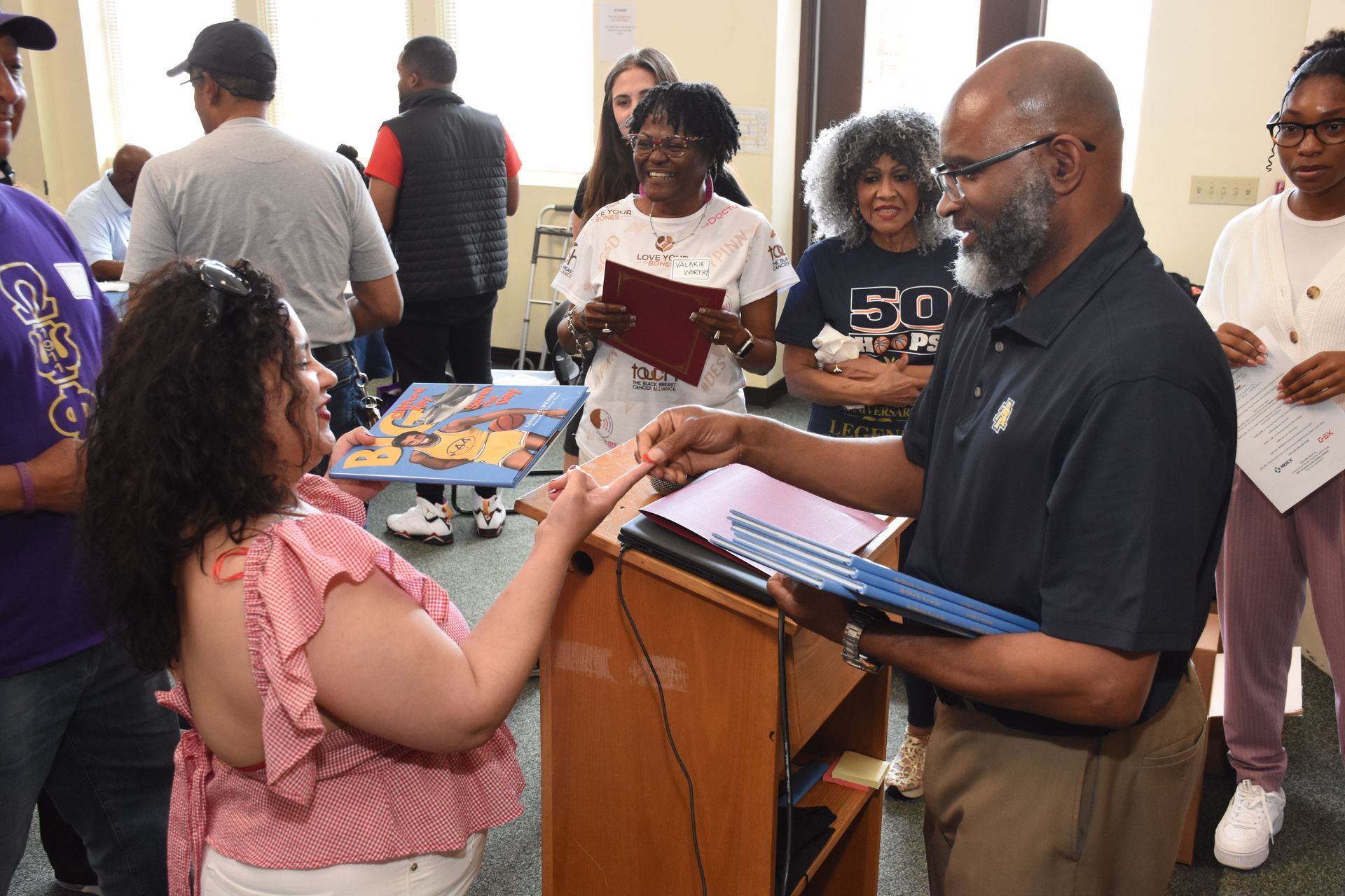 Man handing a certificate to a woman, surrounded by people in a room.