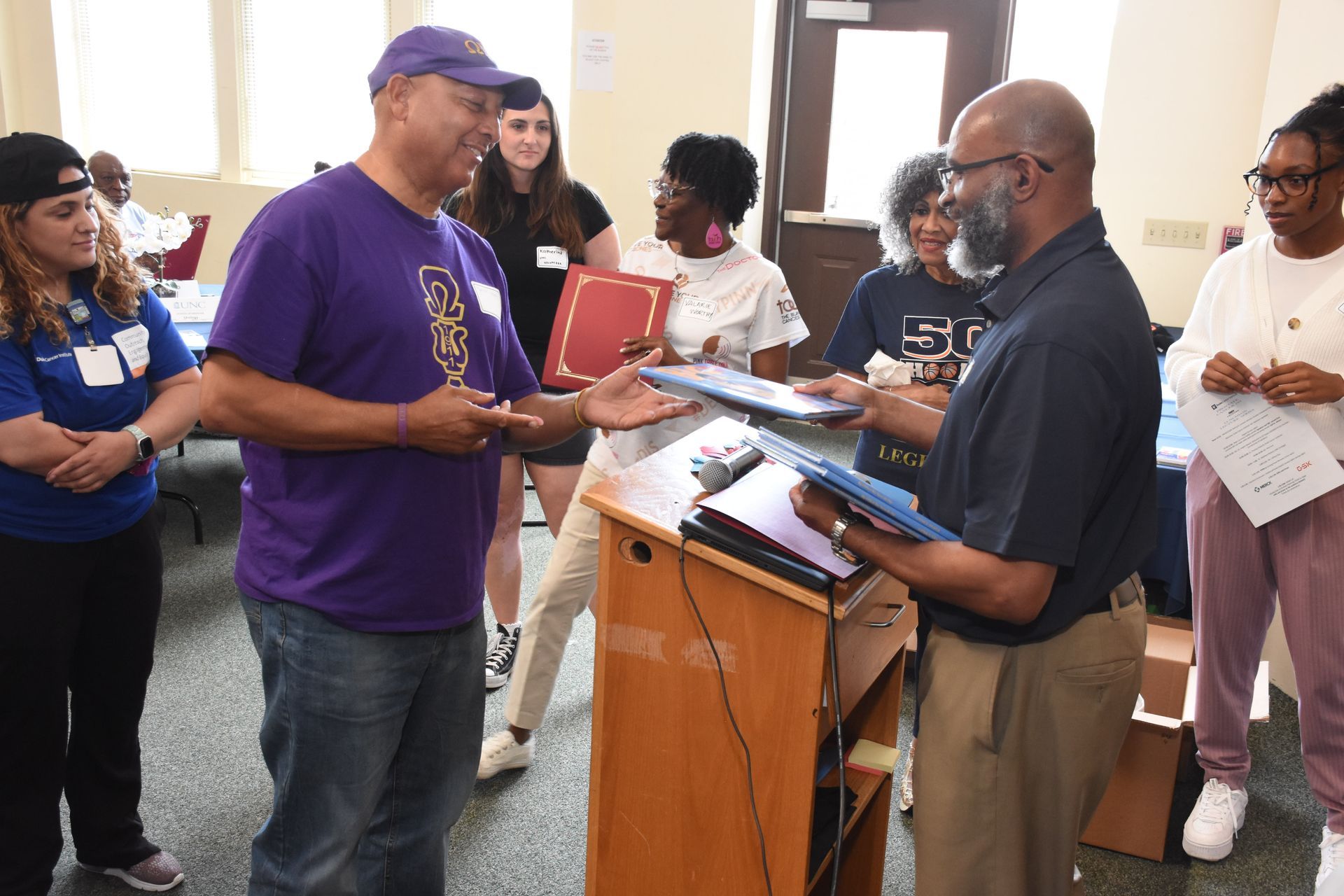Man in purple shirt receiving a certificate from another man at a podium. People watch.