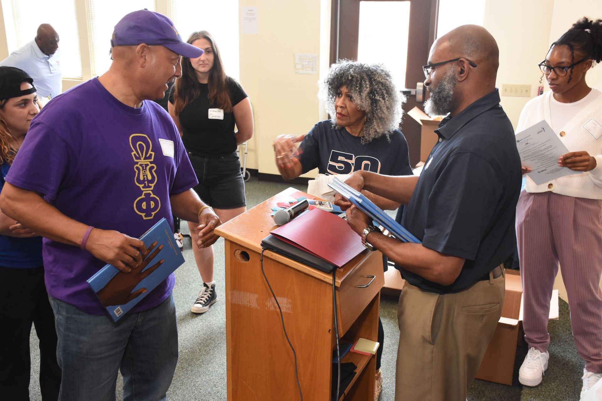 People at a podium, exchanging documents. A man in purple cap speaks to another man. Others watch.