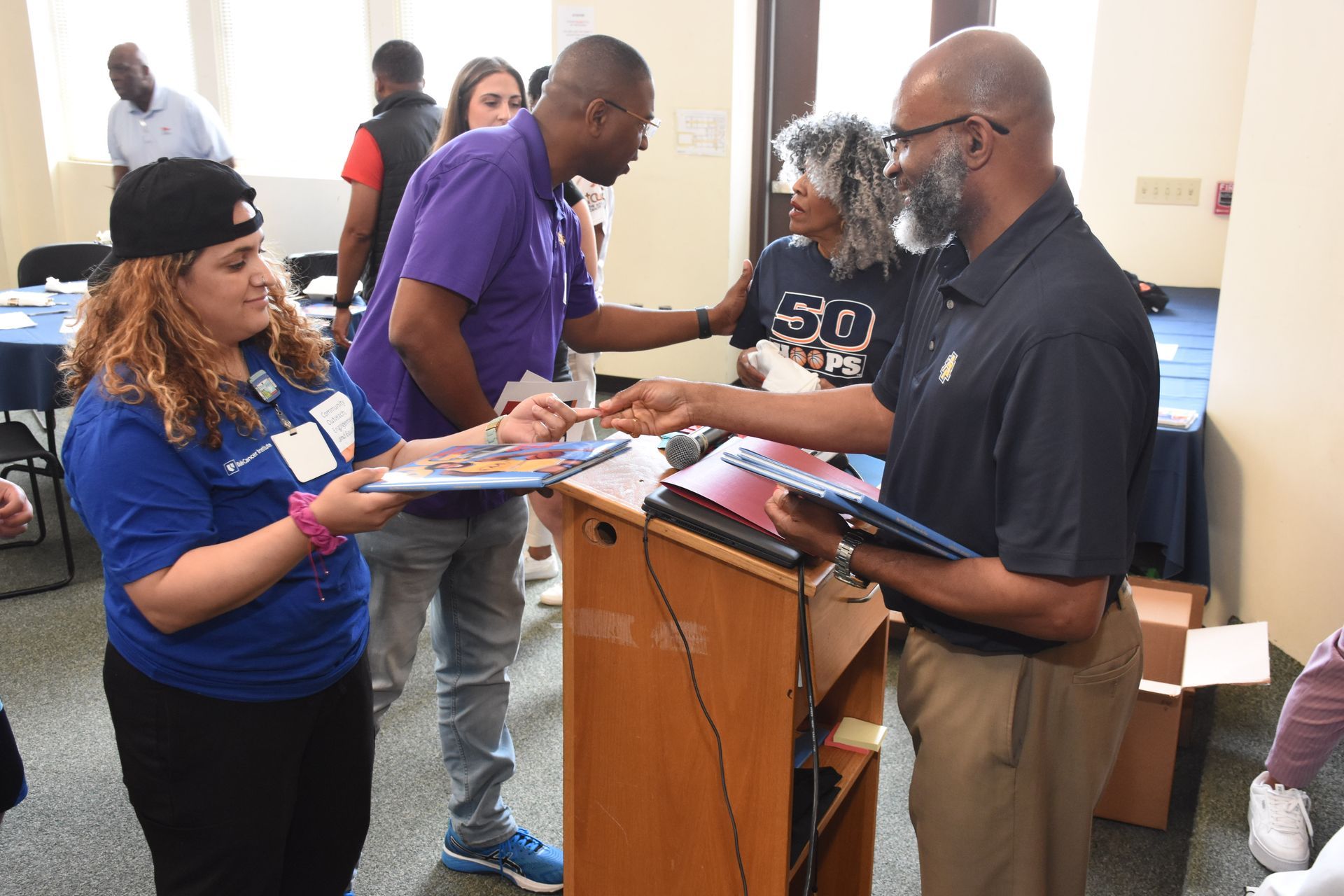 People passing out folders in a room, some wearing blue and purple shirts.