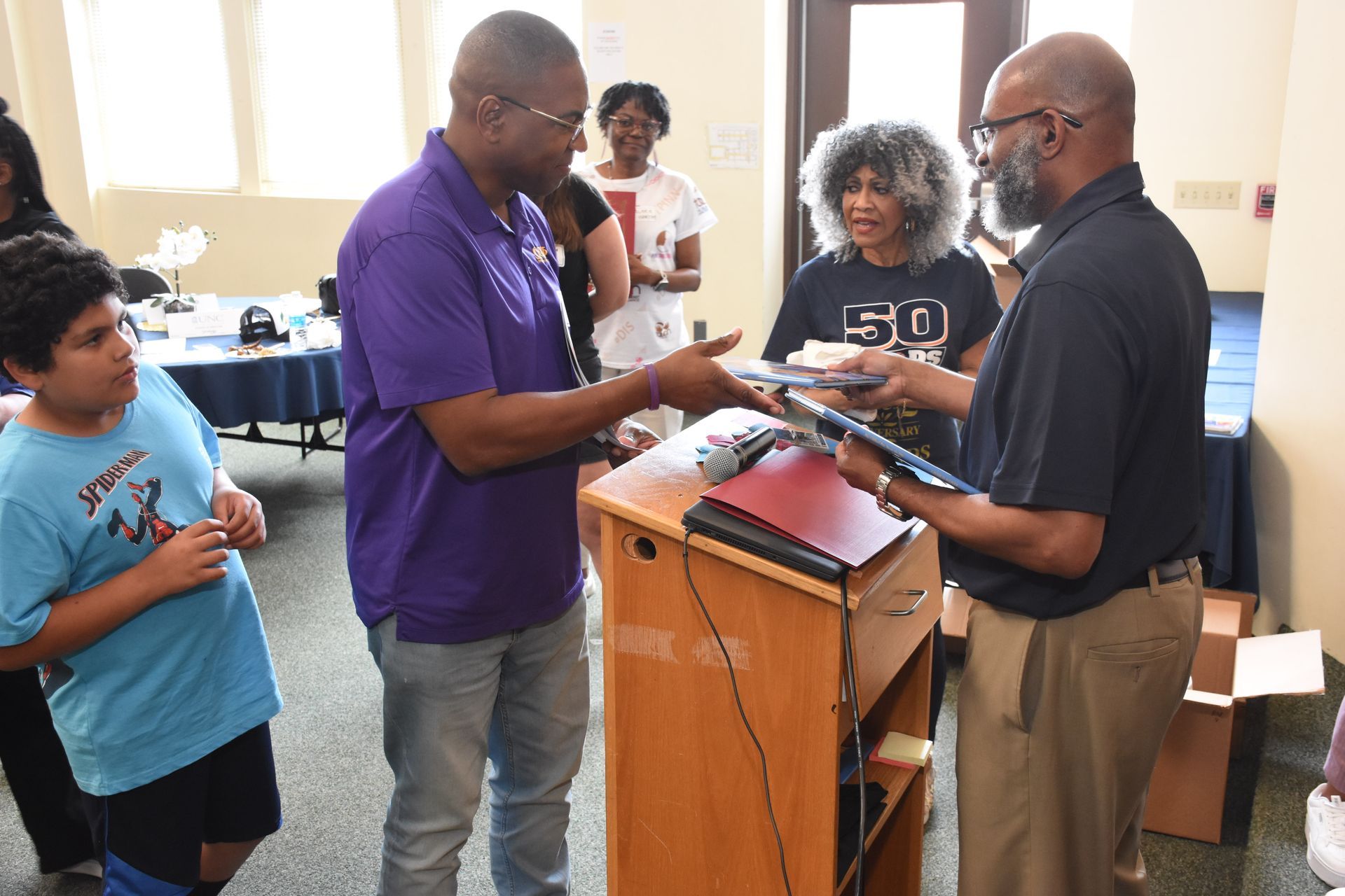 Man in purple shirt hands laptop to another man. Others look on in a room.