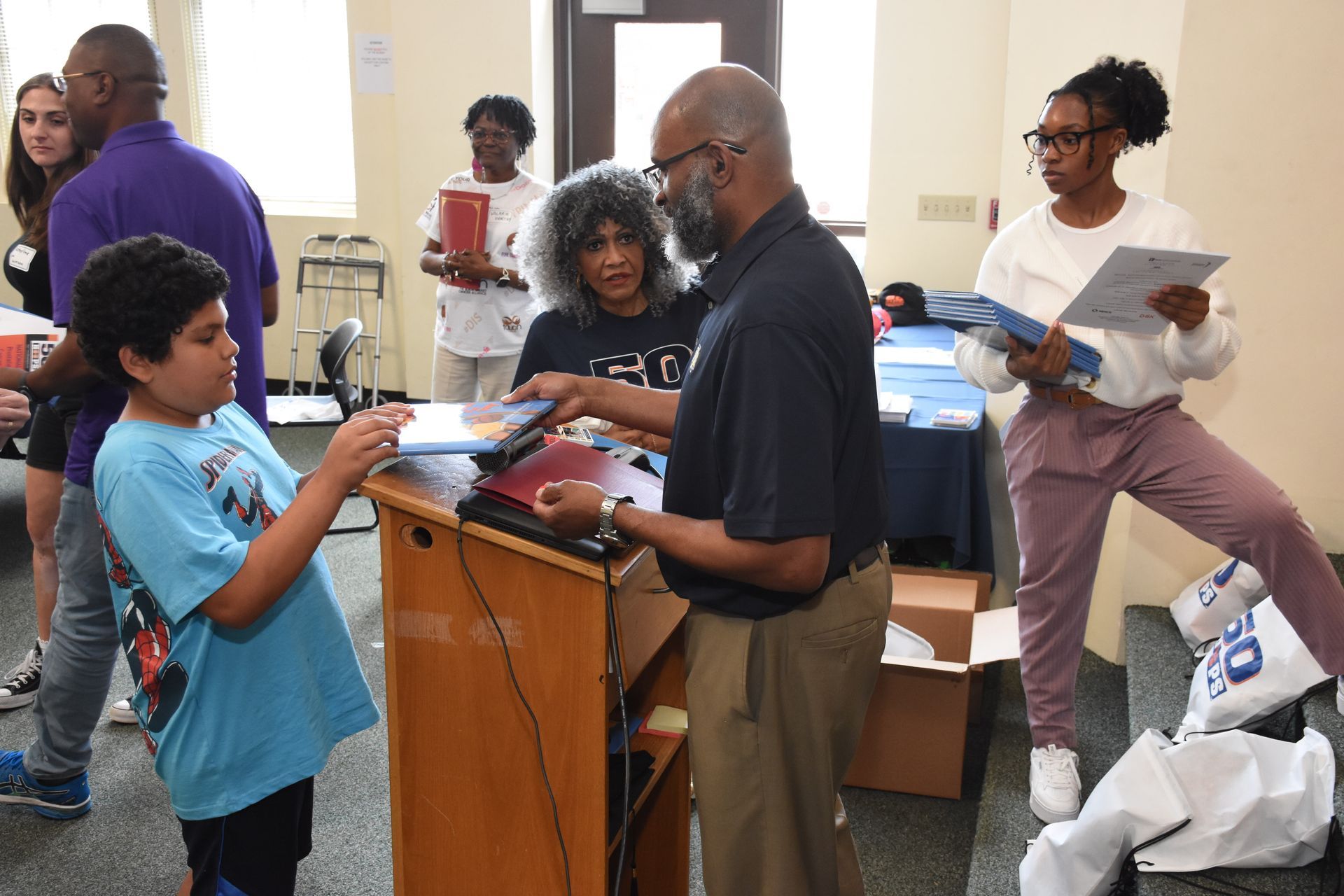 People distributing items at a desk. A child receives an item from a person, others hold papers. Interior setting.