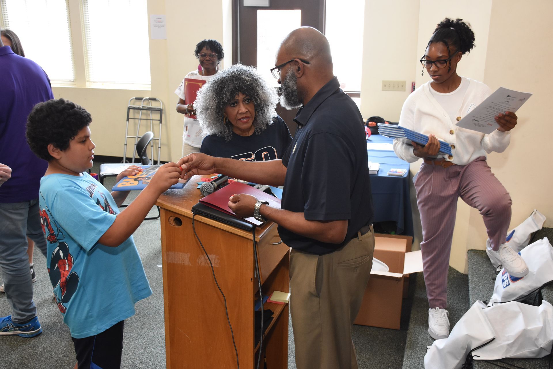 A person hands a gift to a boy at a podium. People in a room observe.