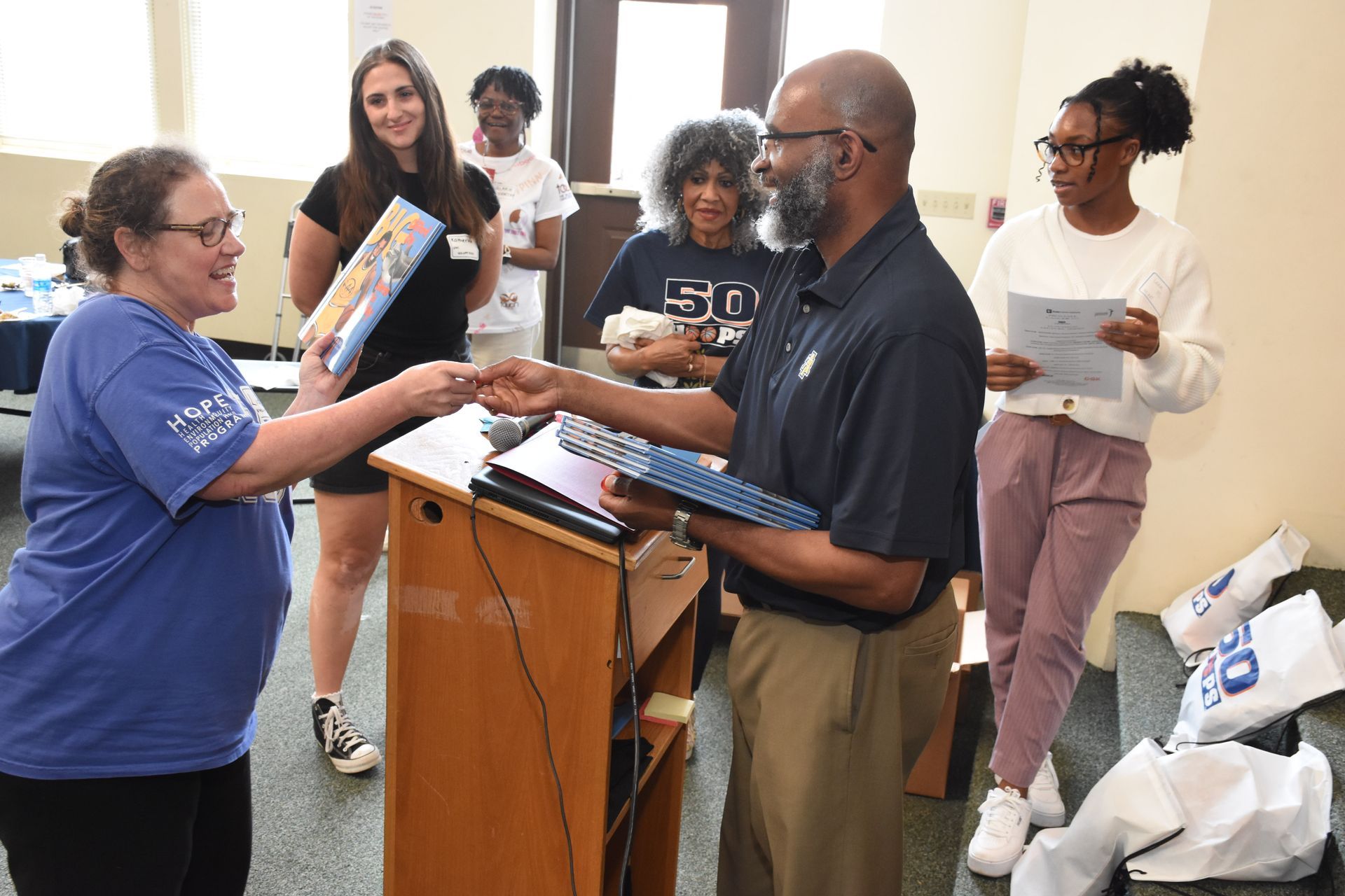 Man presenting a gift to a woman, group of people in background. Indoors, podium, blue and tan clothing.