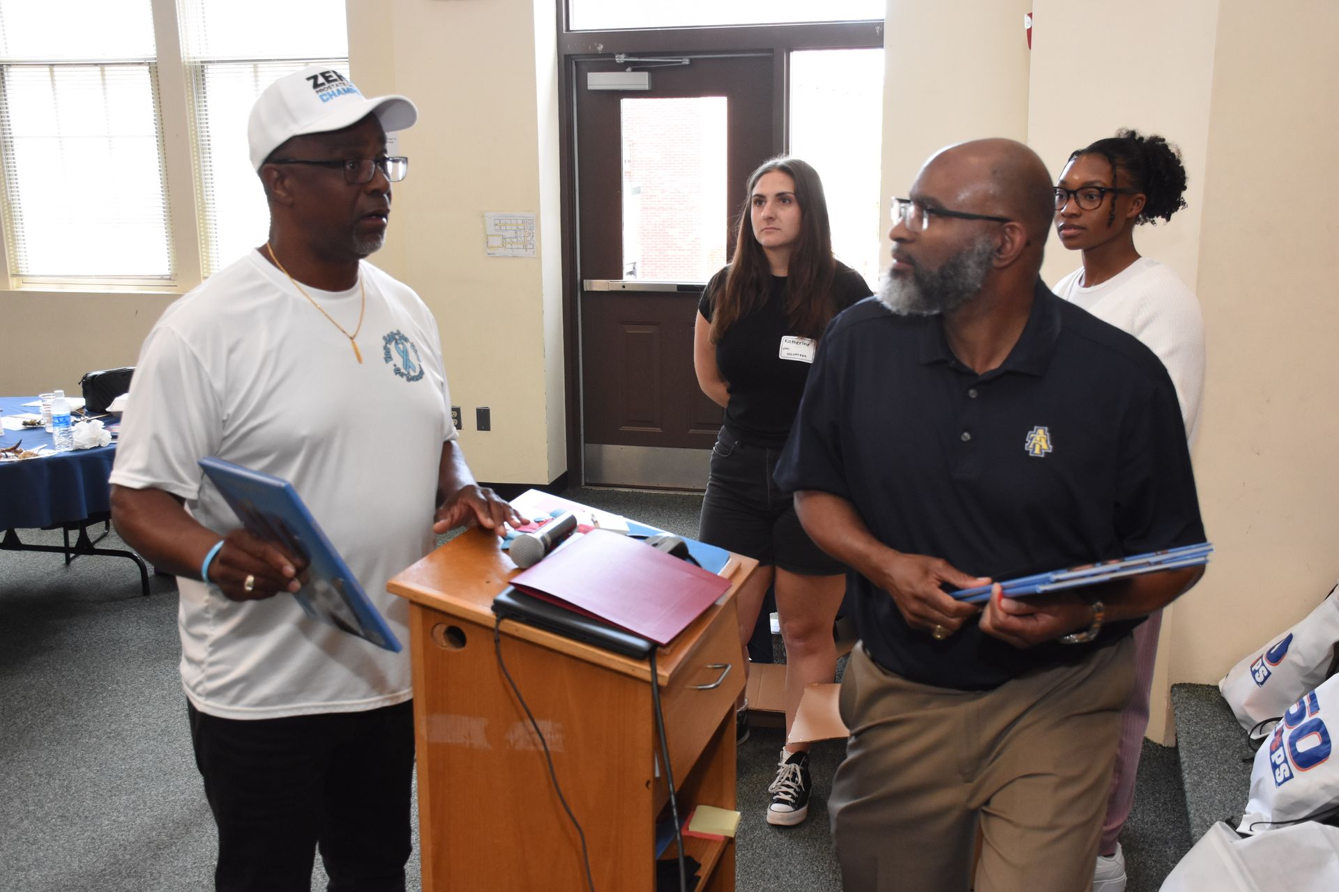 Two men stand at a podium, speaking. Other people in the room listen.