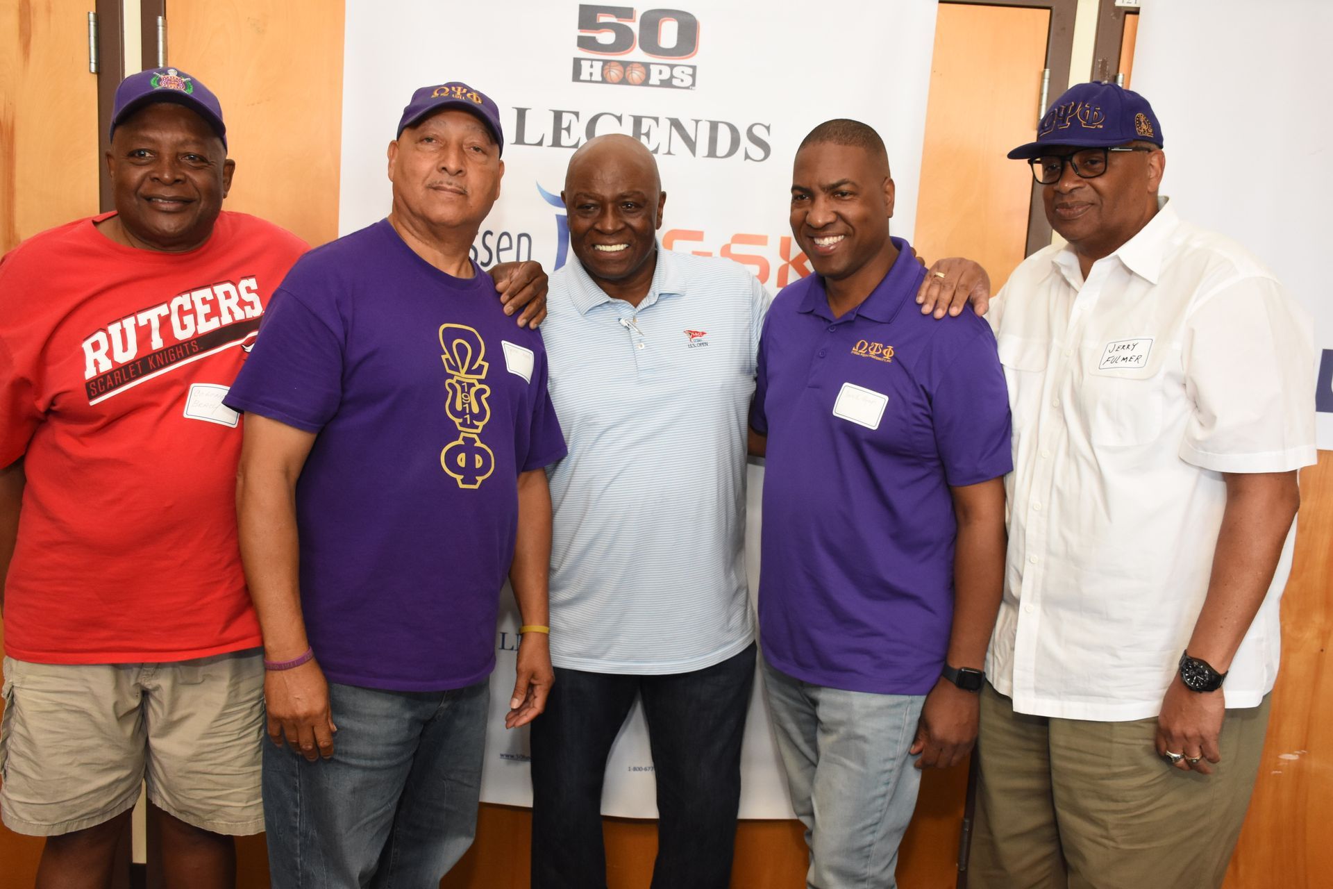 Five men pose together, smiling. Some wear purple shirts, one a red shirt. Background has a sign.