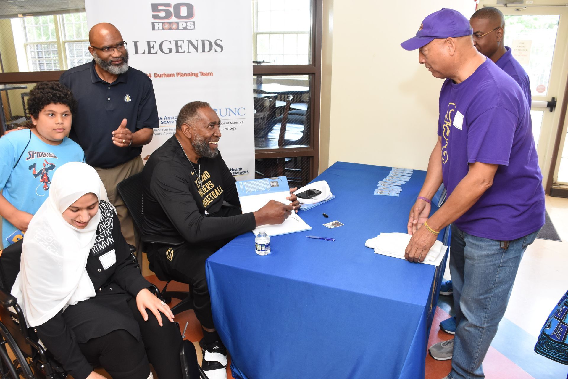 A person signing autographs at a table; others watch. A woman in a wheelchair and a child look on.