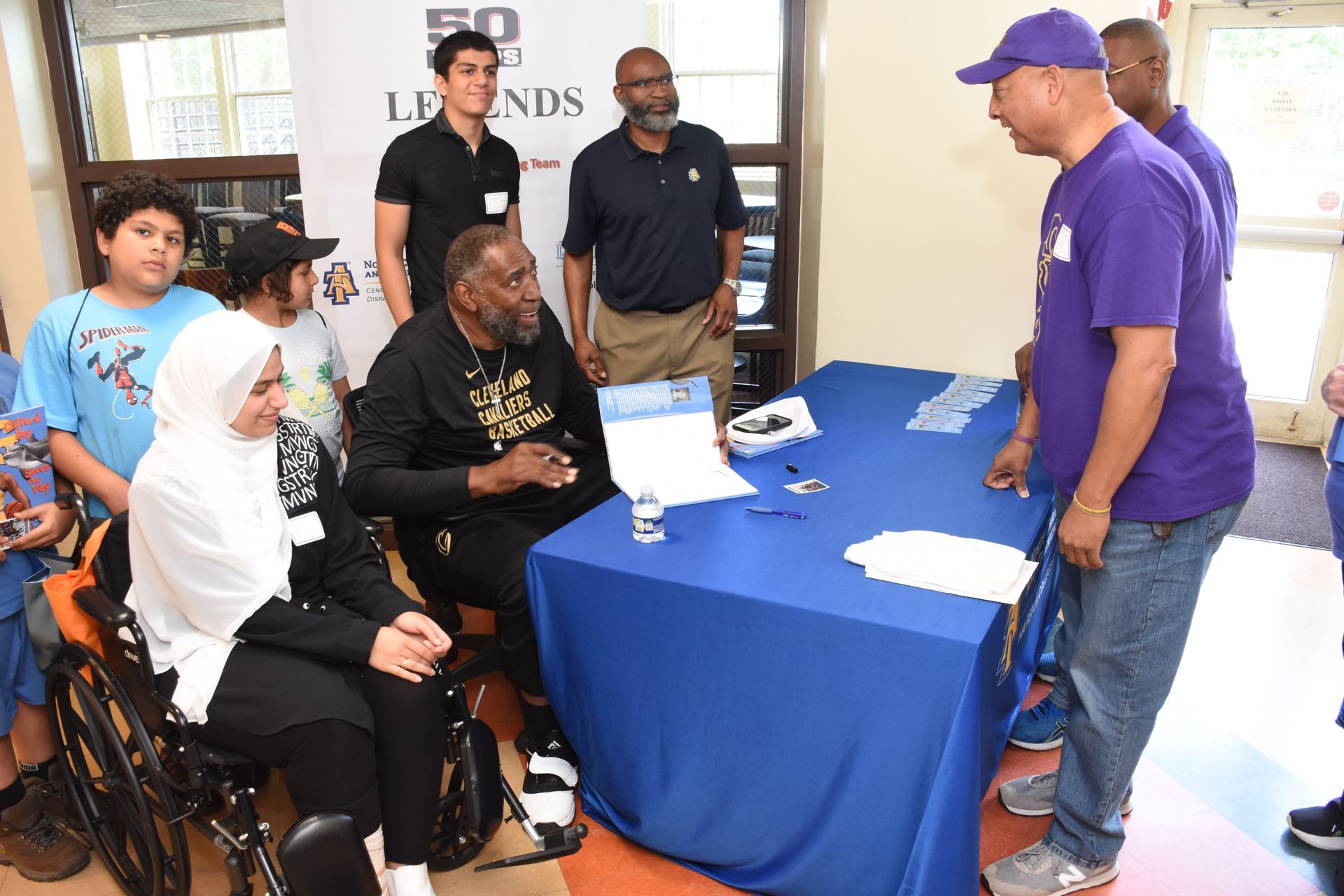 People gather at an event with a table and banner. One person in a wheelchair, others standing.