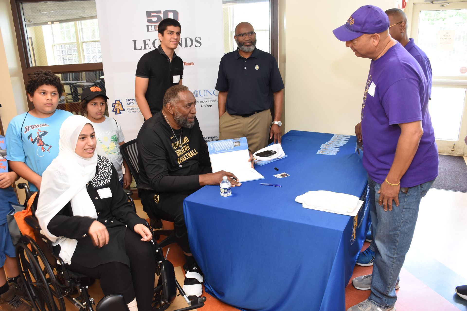 A man signing a paper at a table. Several people observe, including one in a wheelchair and one wearing a purple cap.
