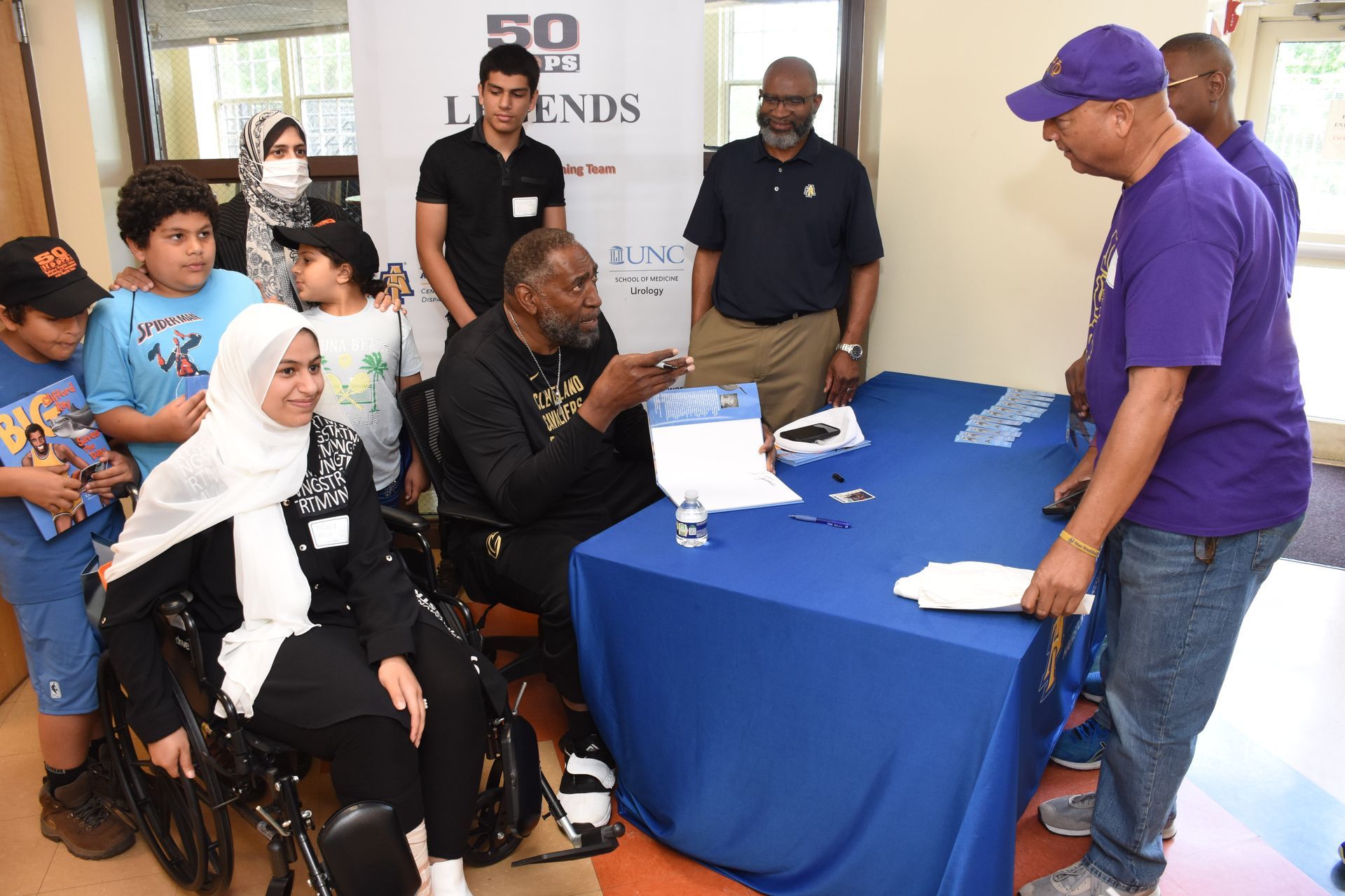 People gather around a table where a man in a wheelchair signs a document. Others watch nearby.