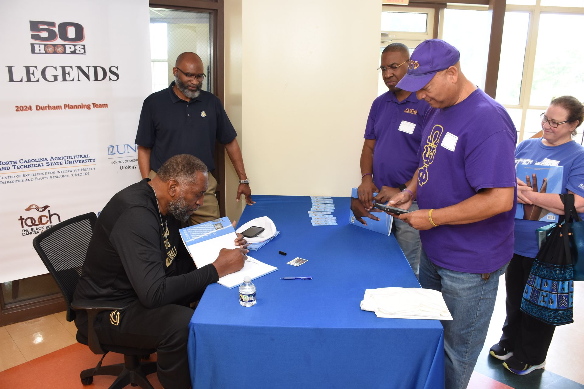 Man signing documents at a blue table, others observe. Sign reads 