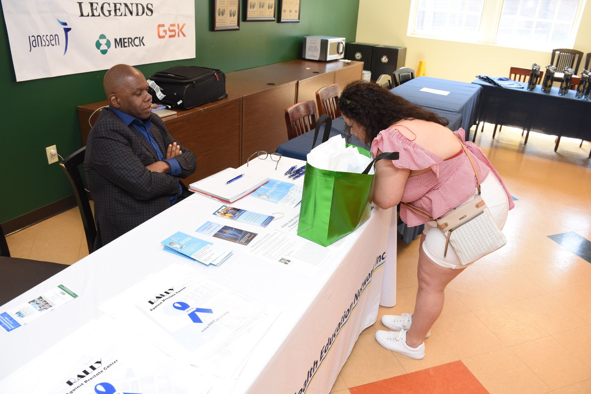 Man at a table with papers, speaking to a person looking at the table. Green bag present.
