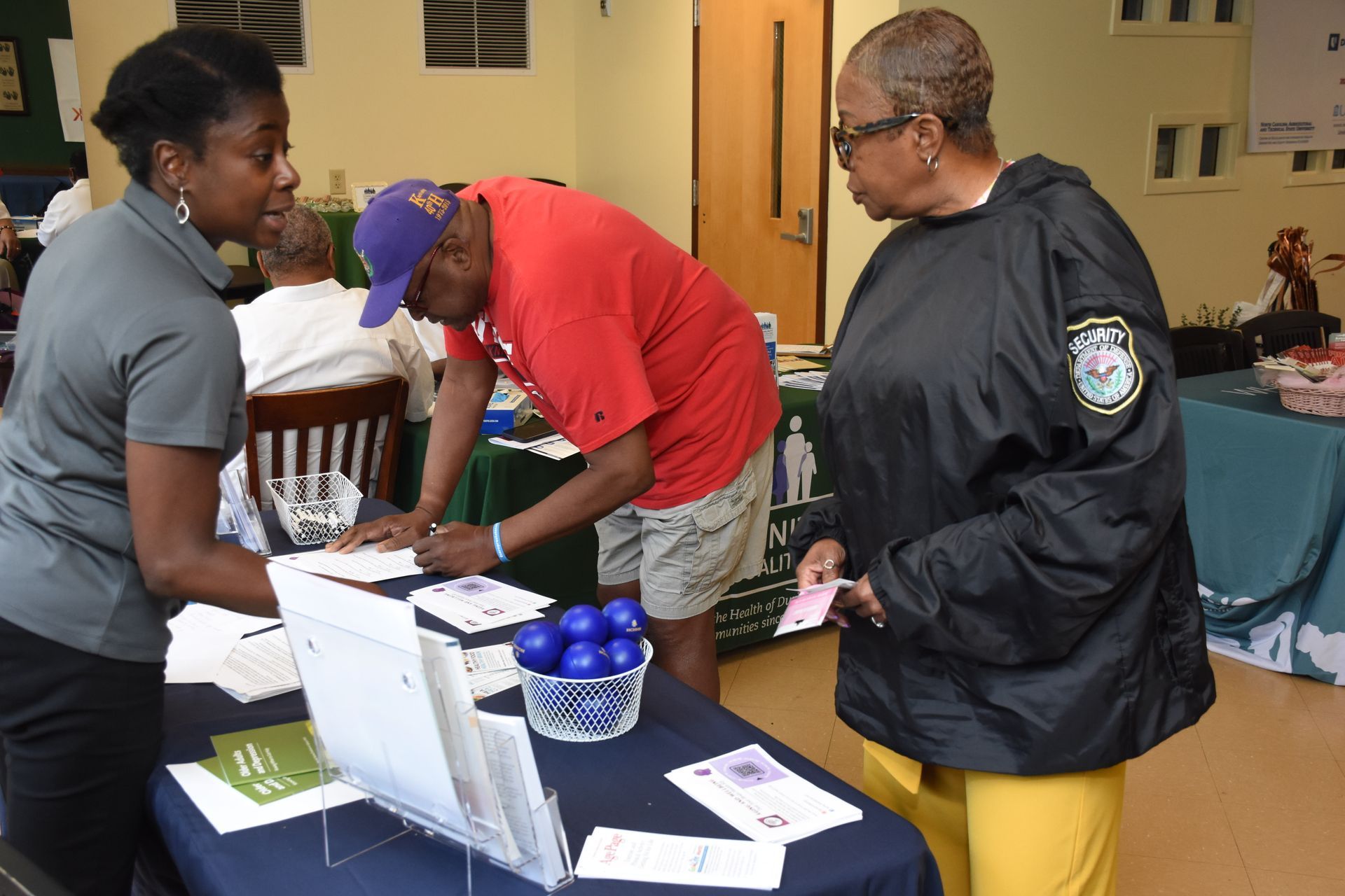 Woman assists two people at a table; one signing a document, another holding paperwork. Indoors.