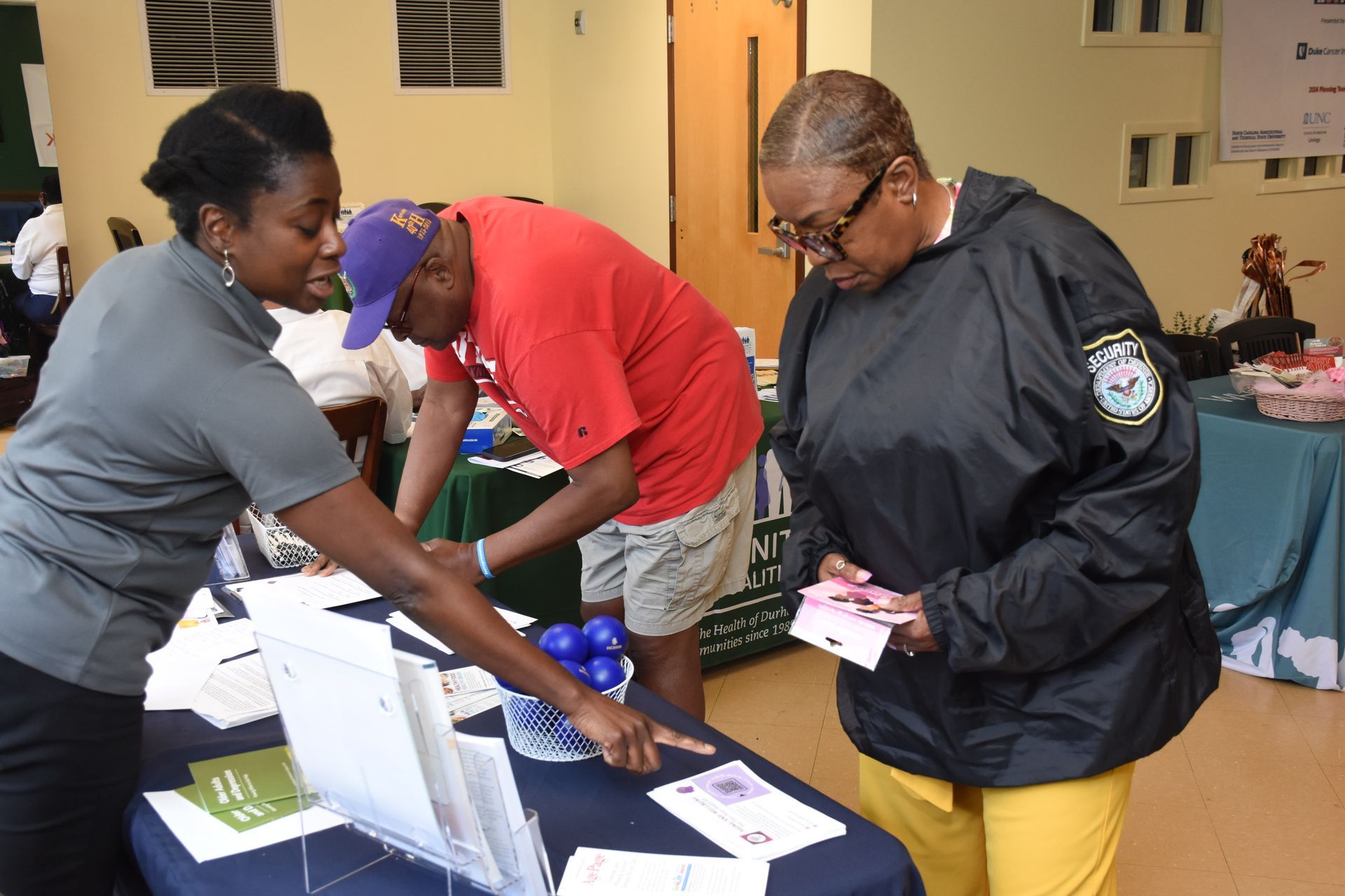 Woman at table assisting two people, pointing at documents. Blue table, various brochures, indoor setting.