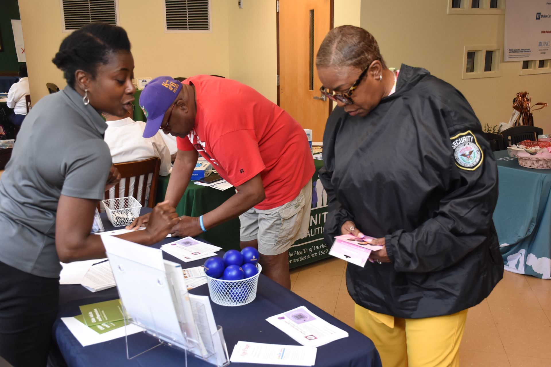 Three people at a table; one shows a card, one has their arm checked, and one looks on.