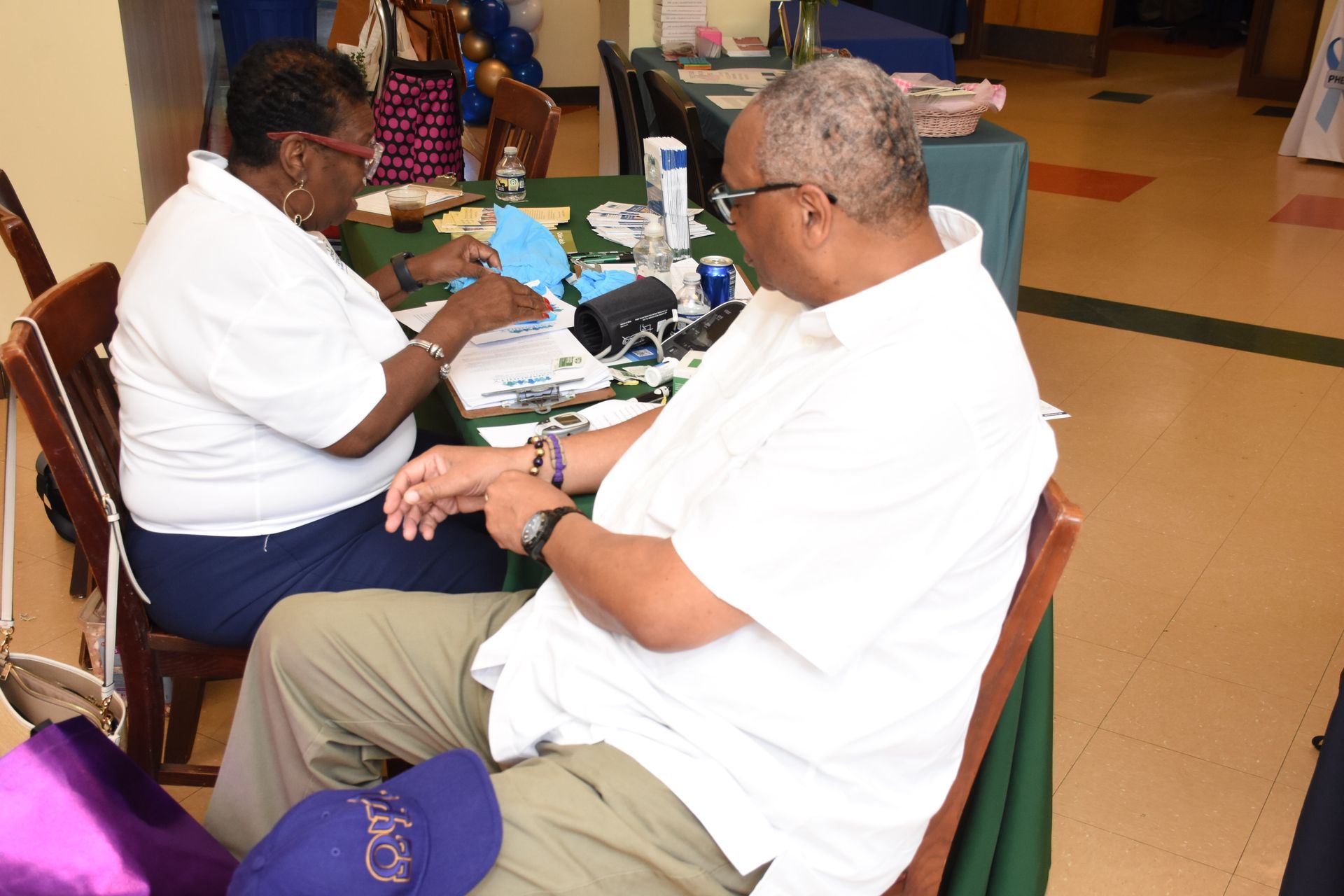 Two people at a table, one taking notes. Man wearing glasses examines his wrist. Table has items on it.