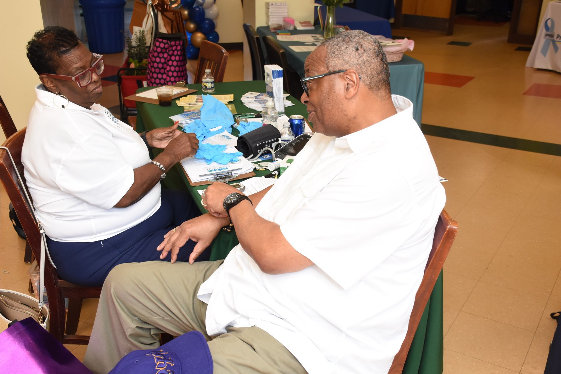 Two people sit at a table, discussing items. One person touches their arm.