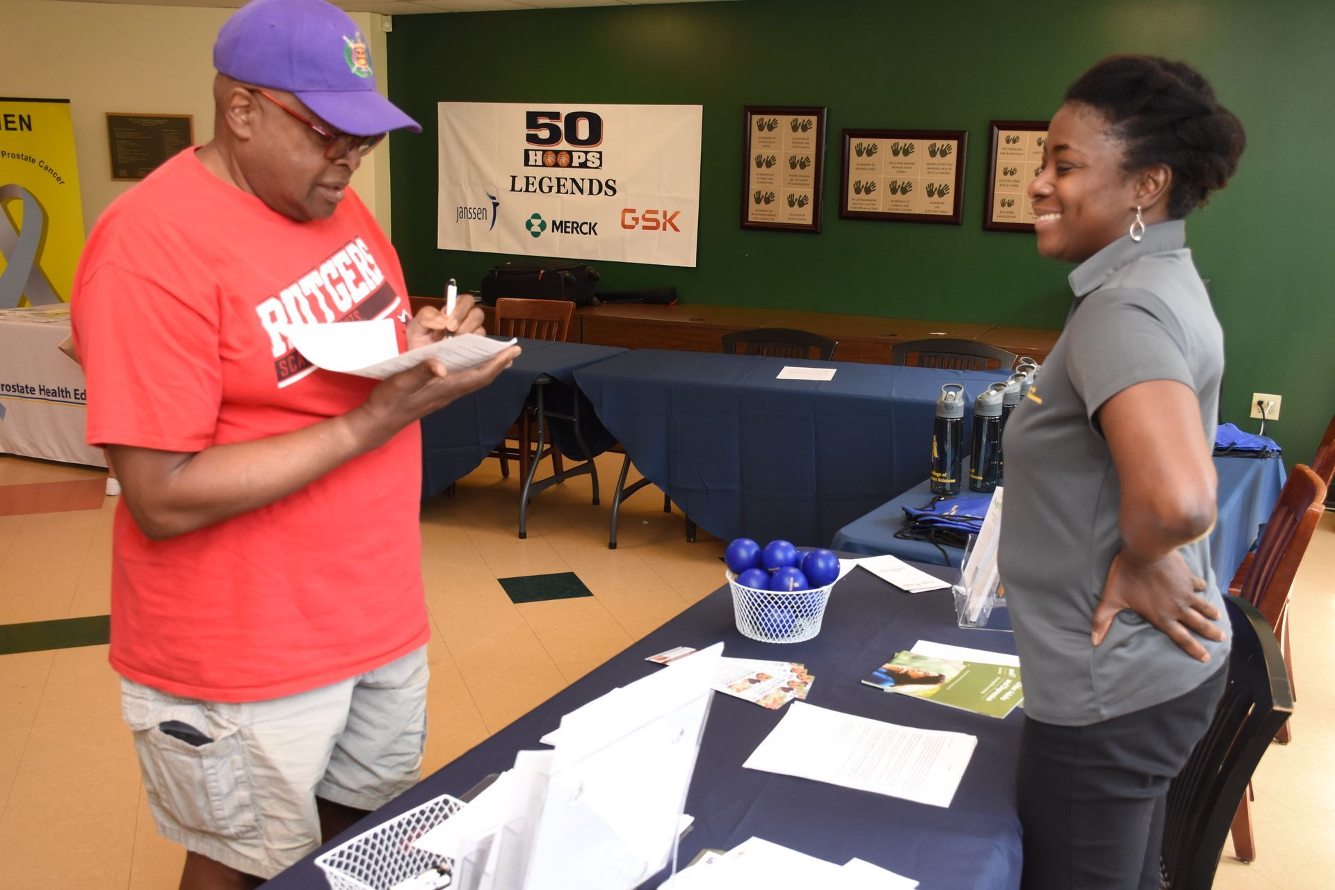 A man in a purple hat and red shirt signs a paper at a table with a woman in a grey shirt.