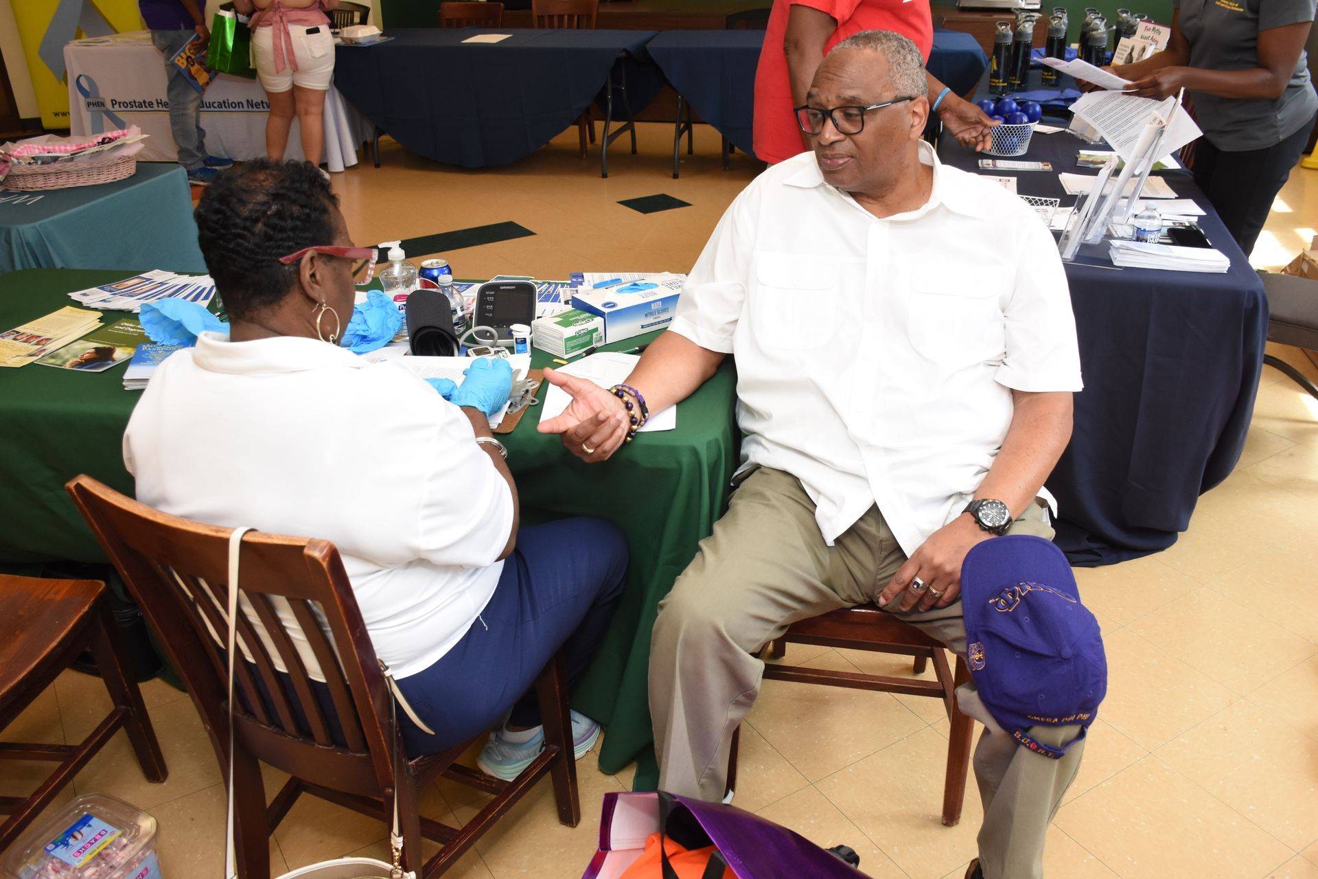 Two people seated at a table, conversing. Man gestures with hand, wearing glasses. Green and blue table coverings visible.