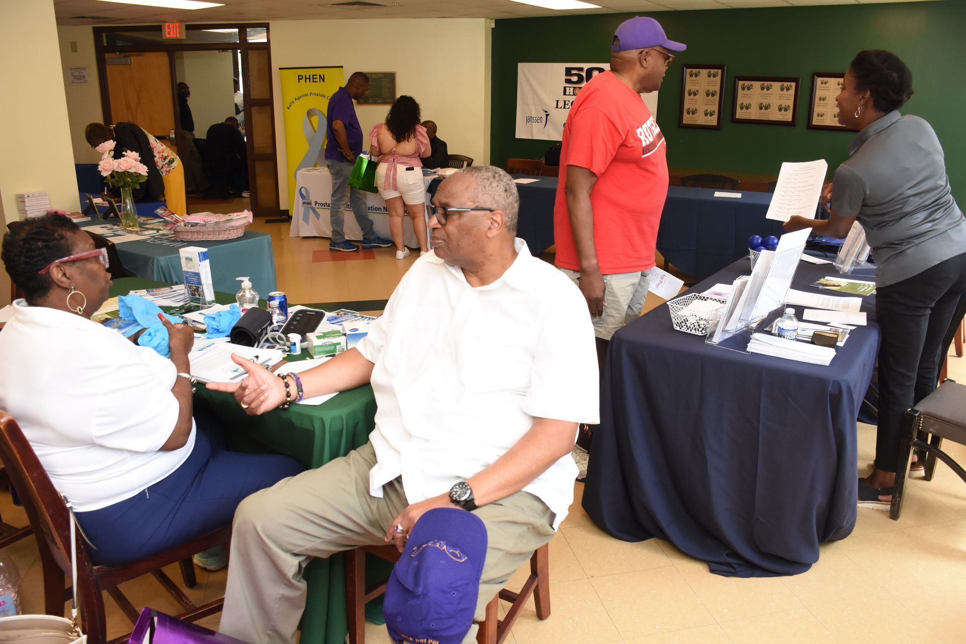 People at health fair, one person's blood pressure being taken. Tables with informational materials.