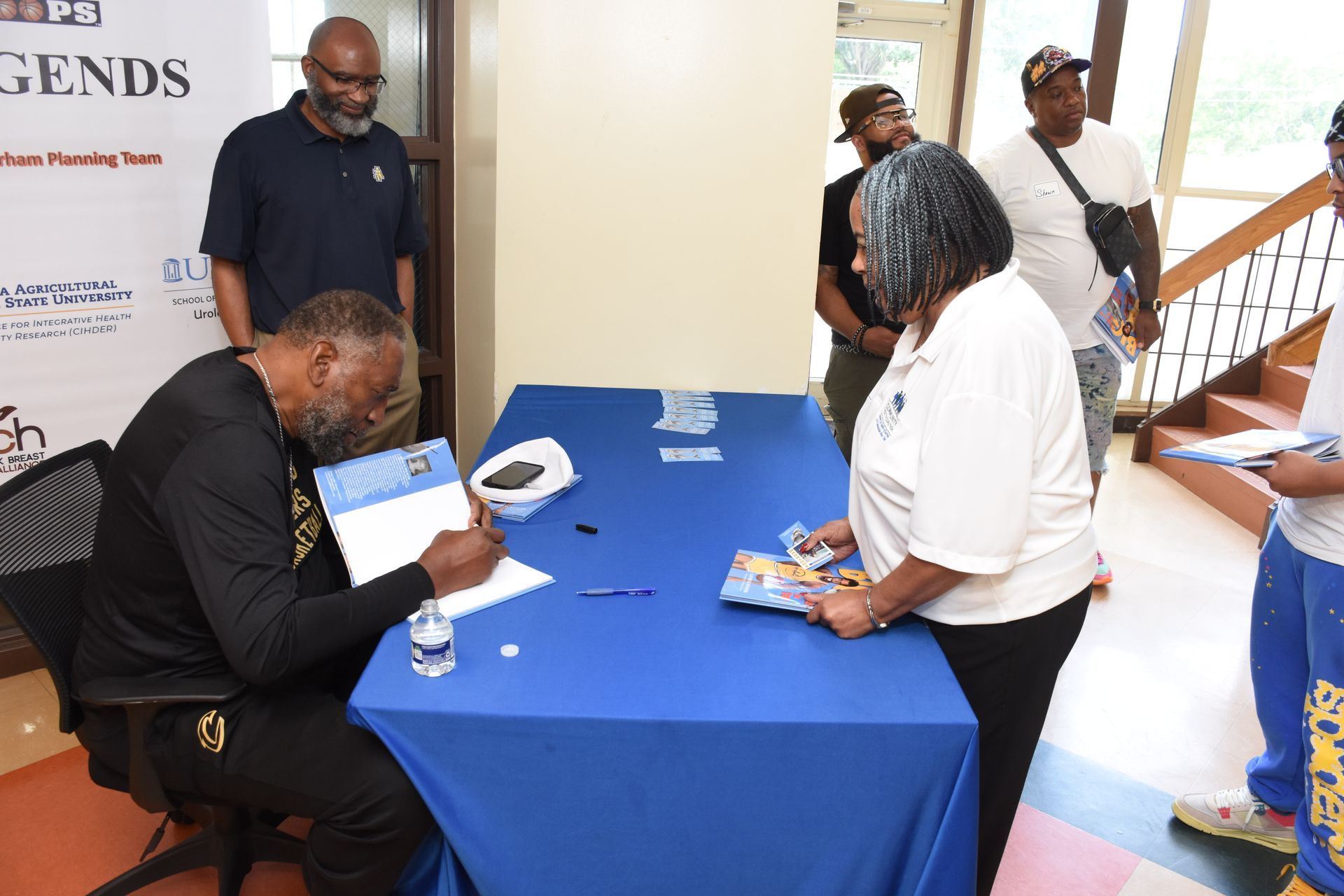 Man signing autographs at a table; people in line to his right. Sign reads 