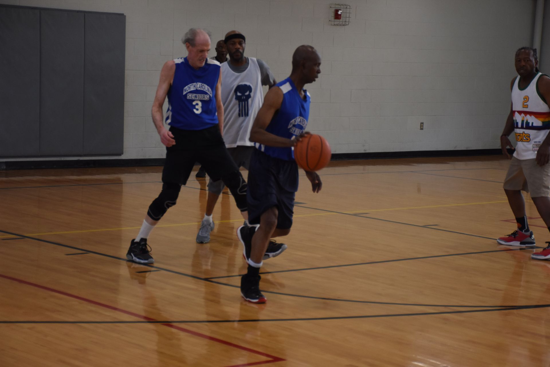 Men playing basketball in a gymnasium; one dribbles, others defend. Wooden floor, blue and white jerseys.
