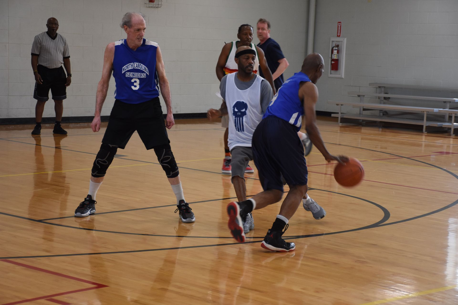 Basketball game in a gym: player dribbles past two defenders. Other players and a referee are visible.