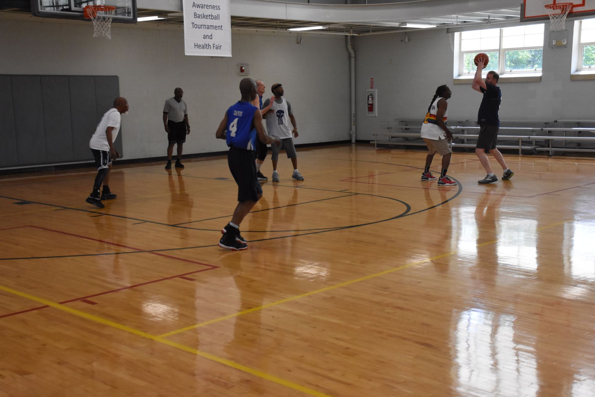 People playing basketball in a gymnasium. One person shoots as others watch. Wooden floor, hoops visible.