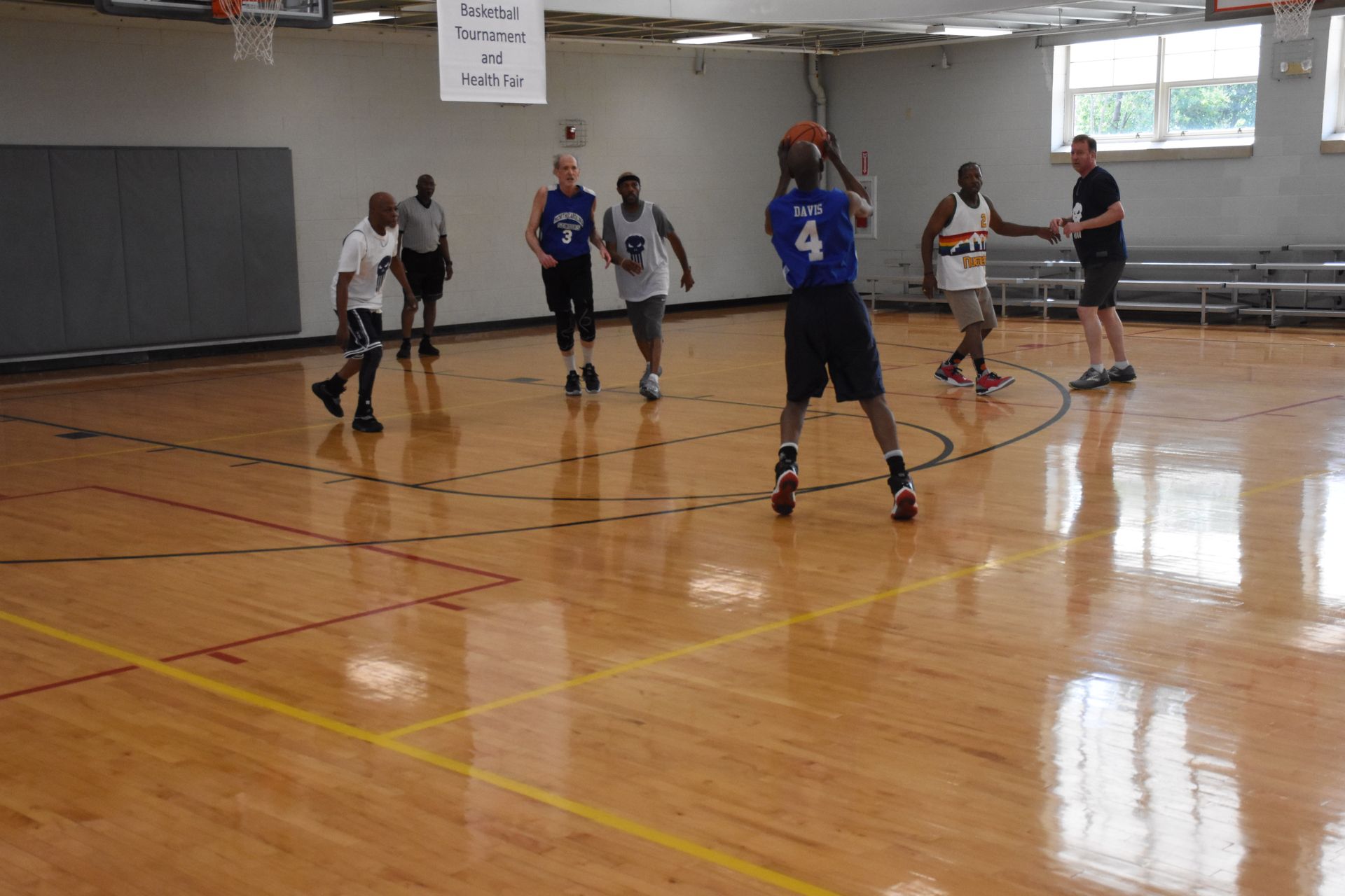 Basketball game in a gym. Player in blue jersey shoots; others watch and move. Wooden floor.