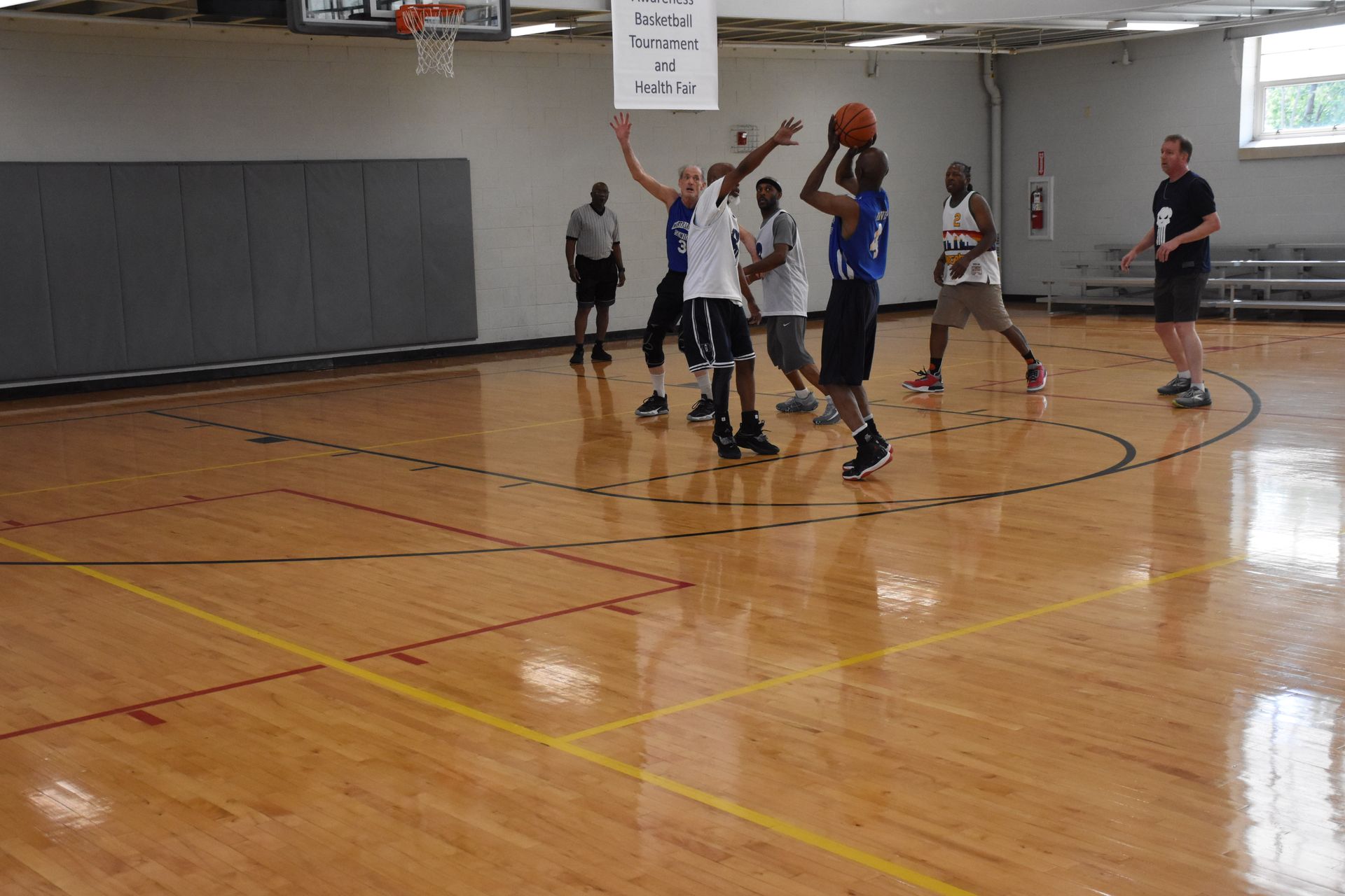 Basketball game in progress: player shooting with defenders, indoors. Wooden court, ceiling lights, spectators nearby.