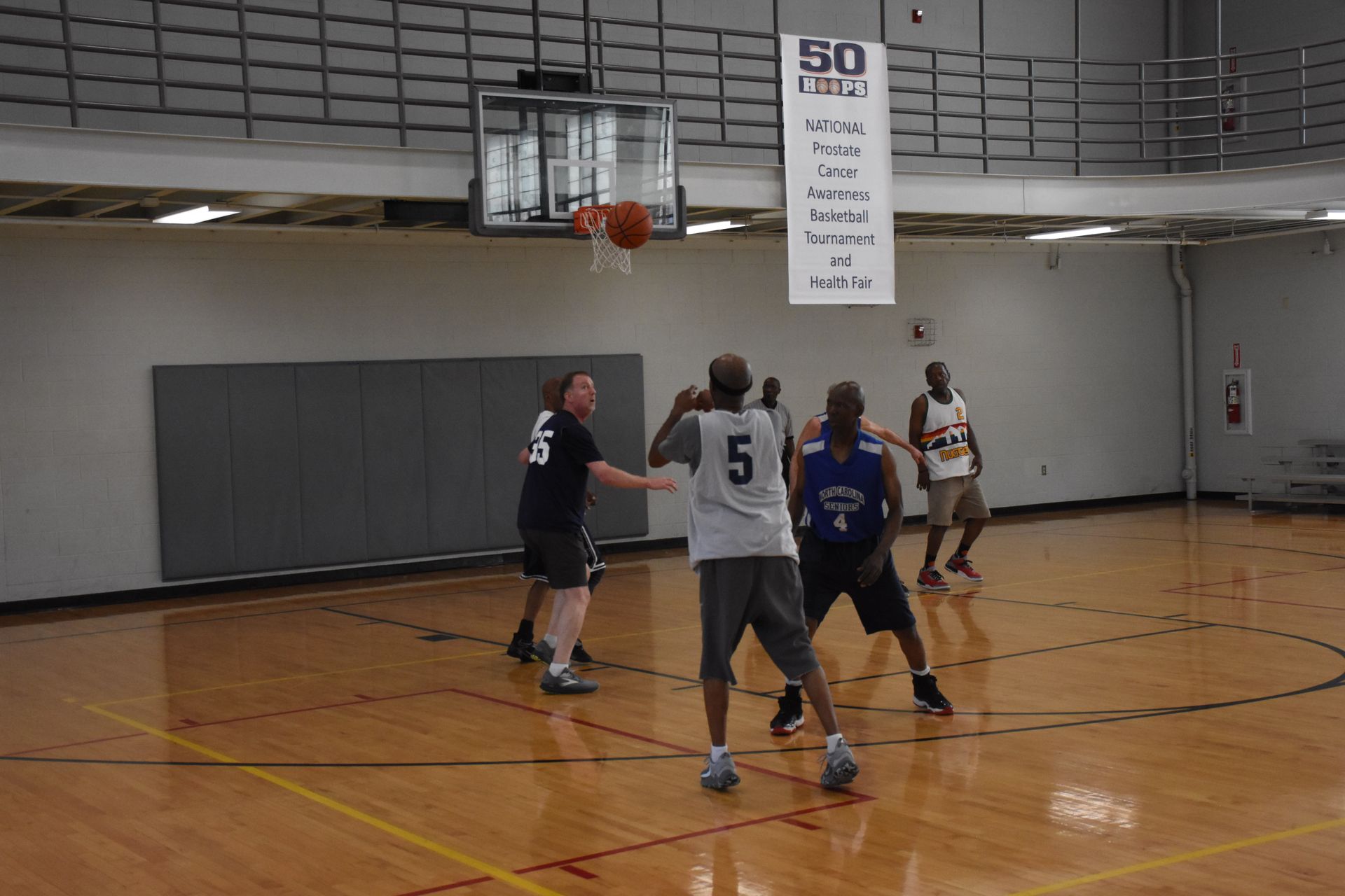 Basketball game in progress: Players on a wooden court, one shooting toward the hoop, others defending.