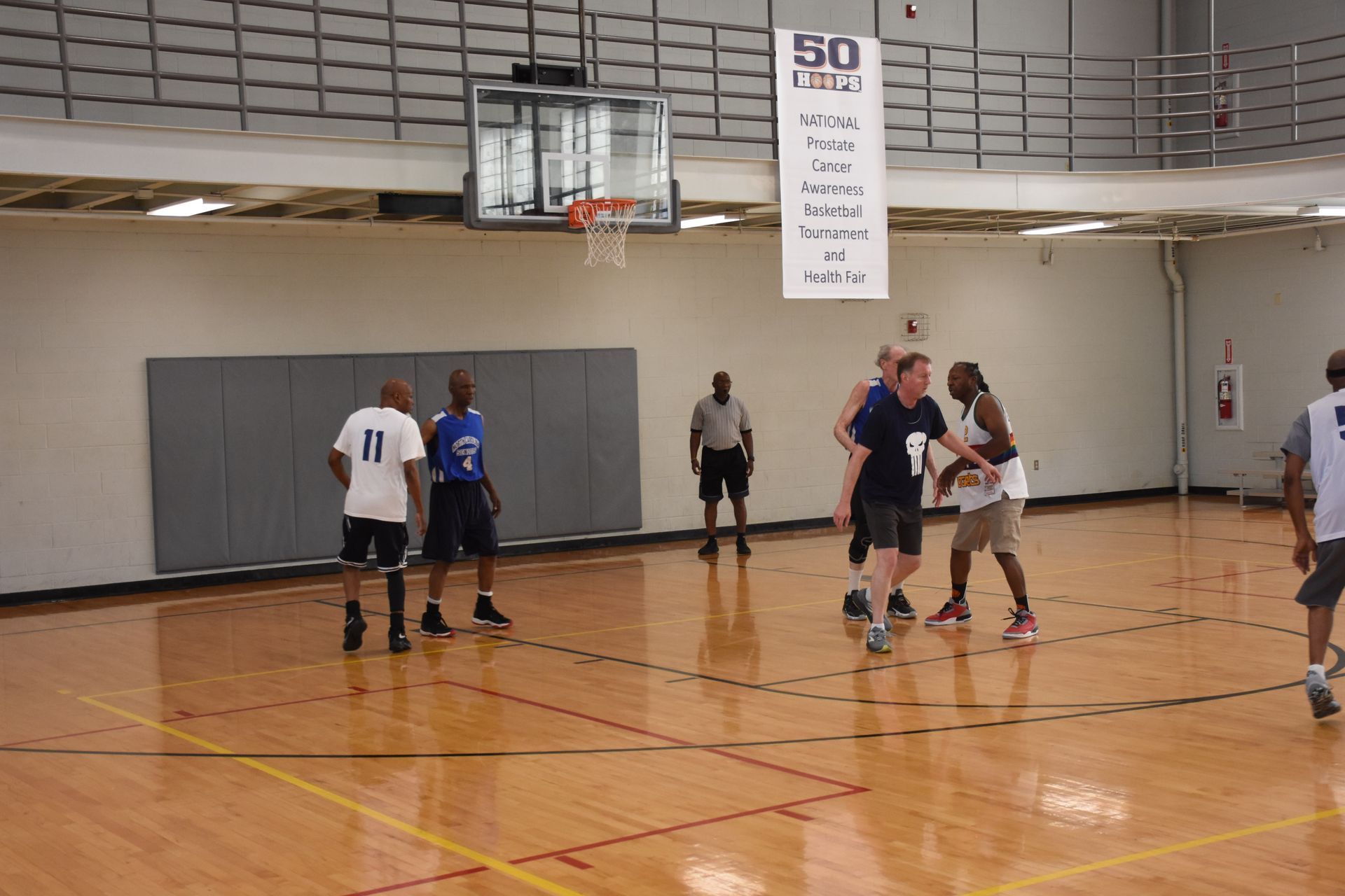 Basketball game in a gym. Players in shorts, some in white, some in blue jerseys. A referee watches.
