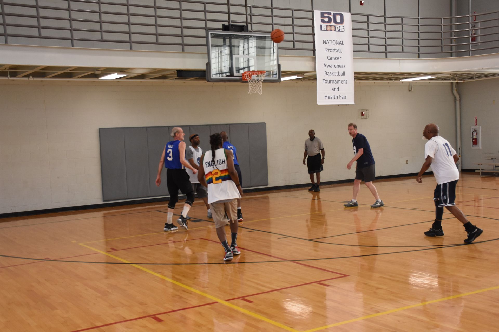 Basketball game in progress: players on court, ball going to basket. Indoor setting with hardwood floor.