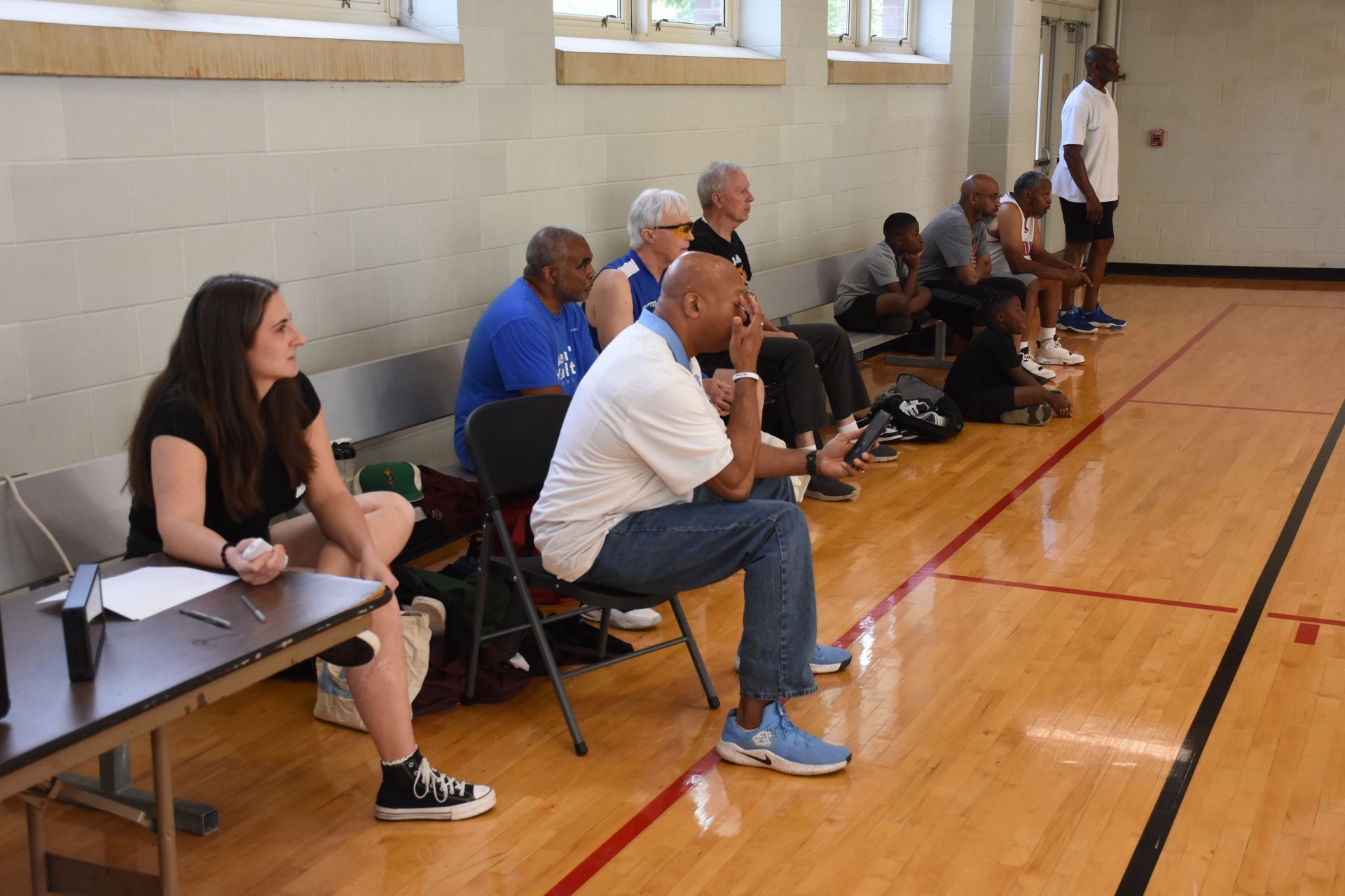 People seated indoors, watching; one woman at a table with a clipboard. Wooden floor and white brick wall.