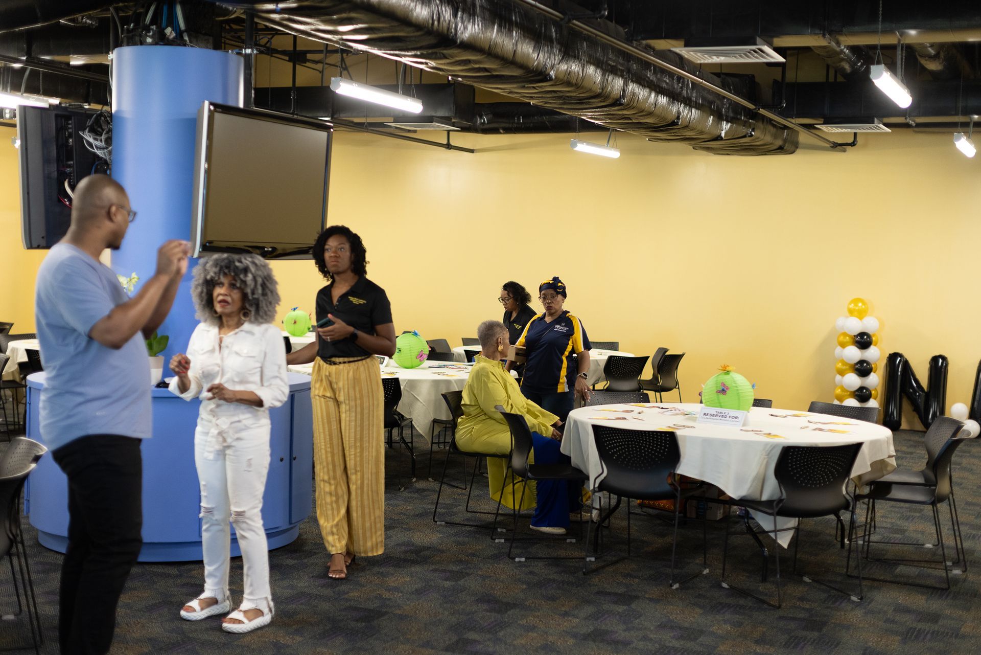 People talking in a room with tables, a blue pillar, and balloons. Yellow and blue colors are prominent.