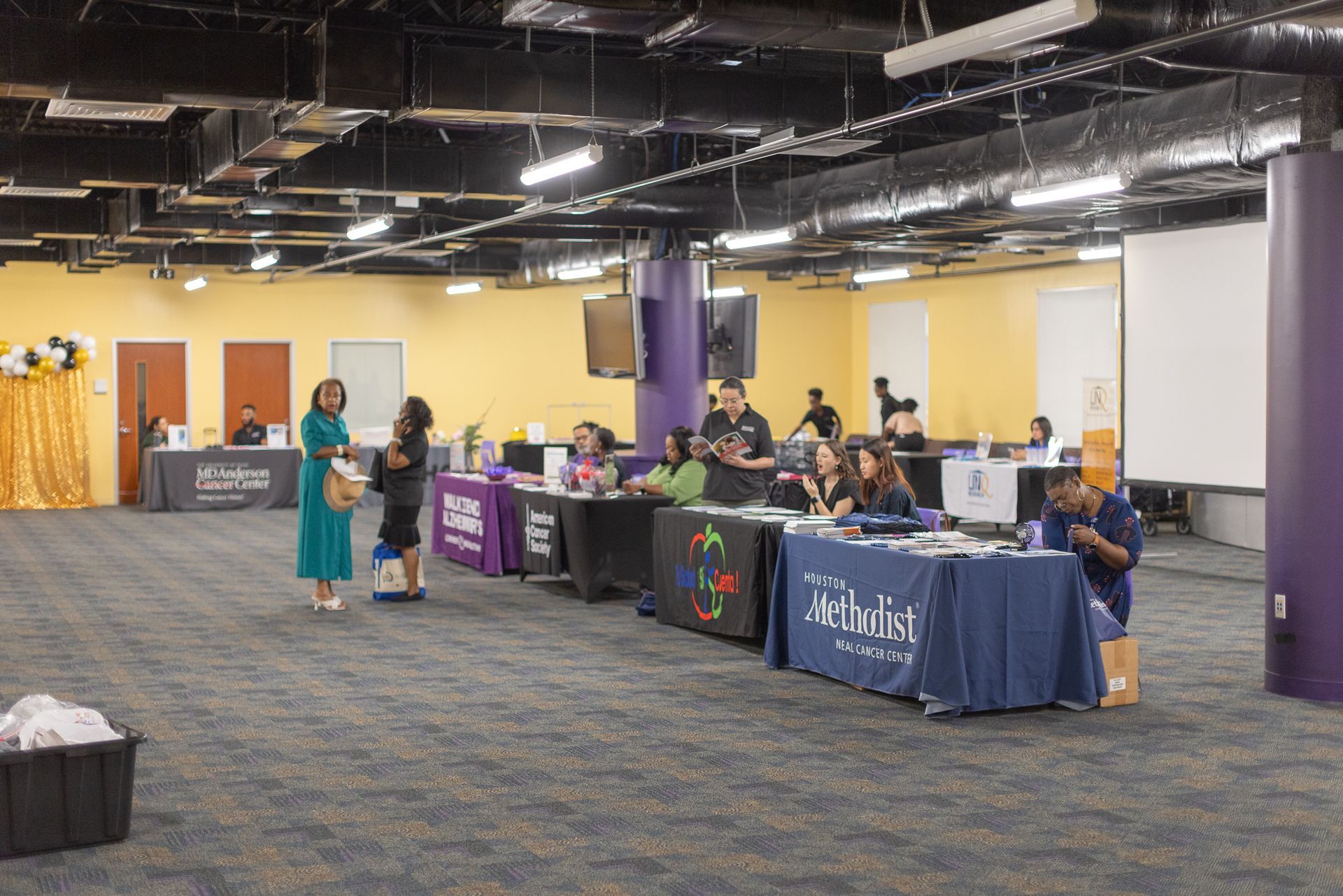 Event with tables, banners, people, and handouts in a large room with purple columns and yellow walls.