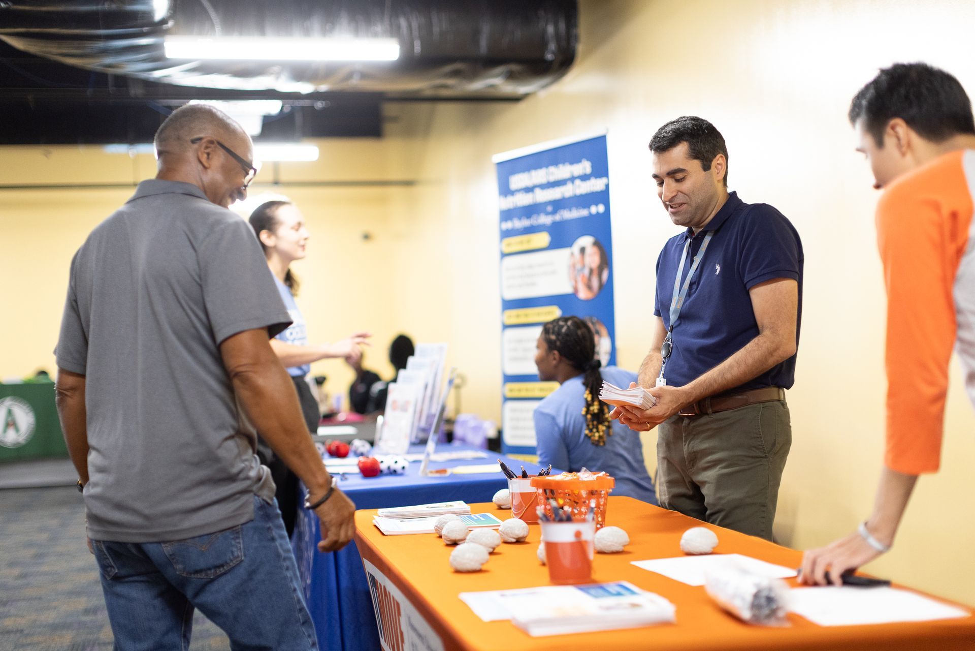 People at an event booth. A man shows objects to two others. Table with pamphlets and display.